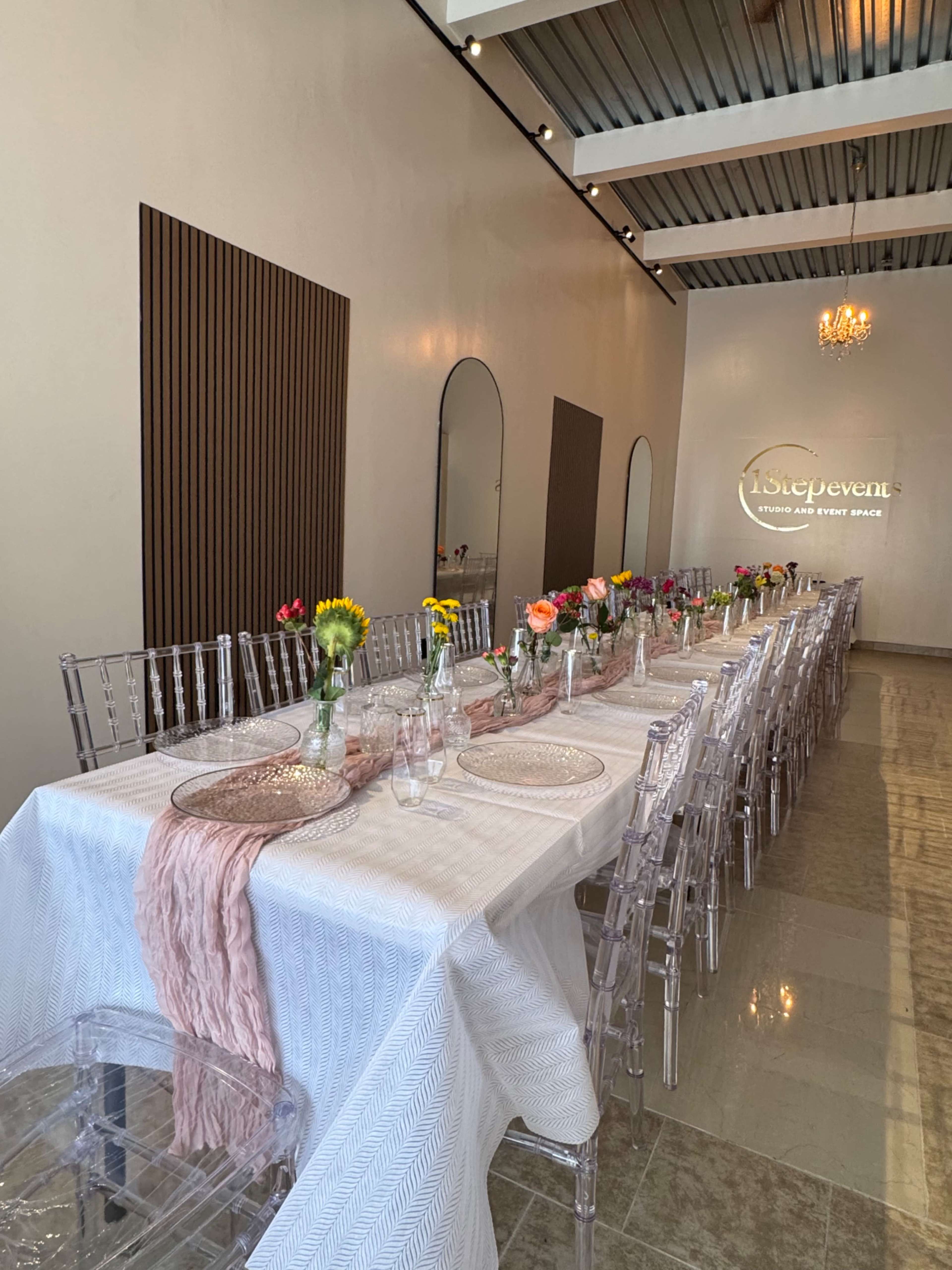 A long, elegantly set dining table with flowers in glass vases is arranged in a well-lit event space featuring a chandelier and modern decor.