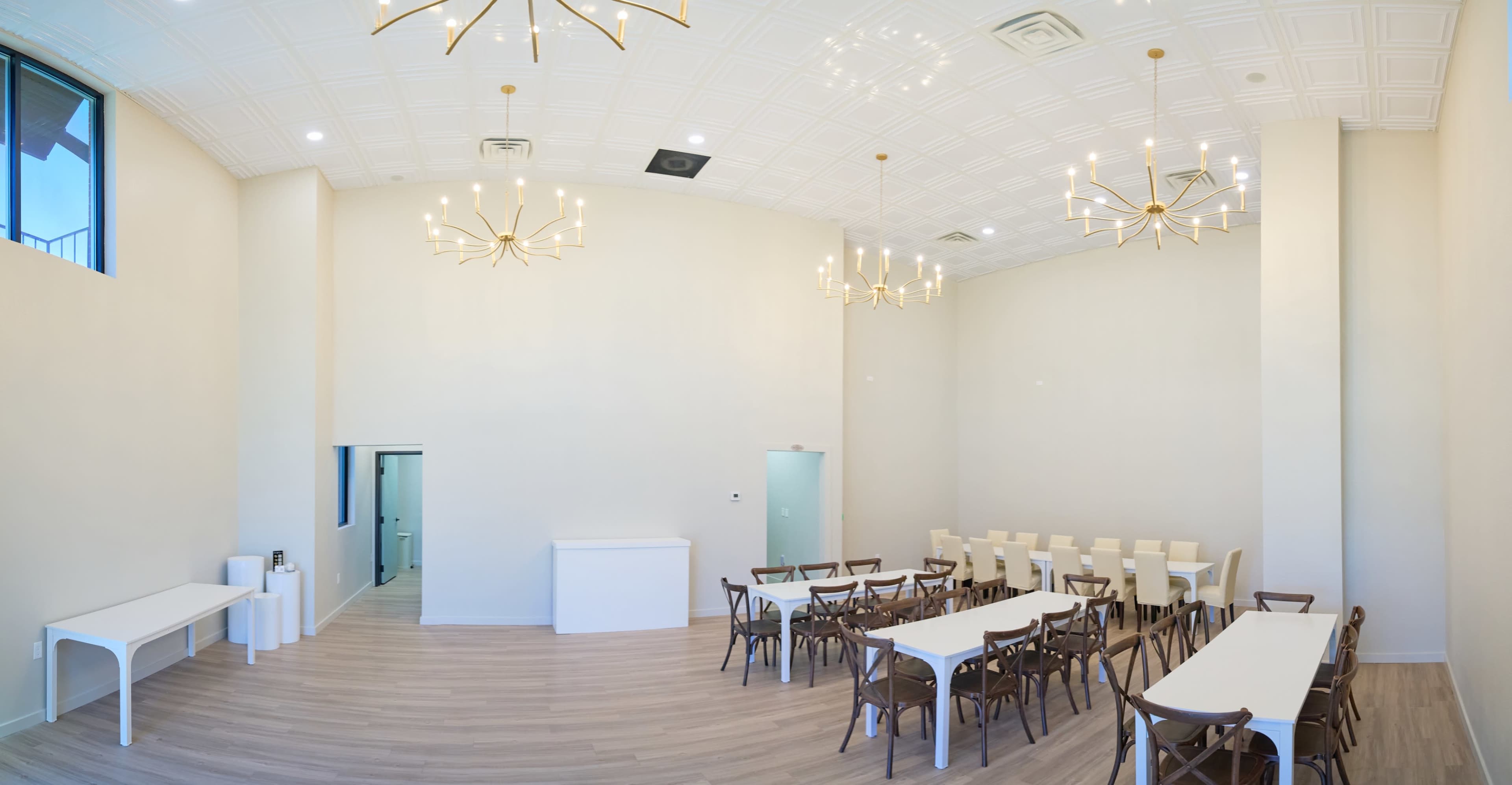 The image depicts a spacious, modern event room with several long tables arranged in a U-shape, surrounded by chairs, and gold chandeliers hanging from the ceiling.