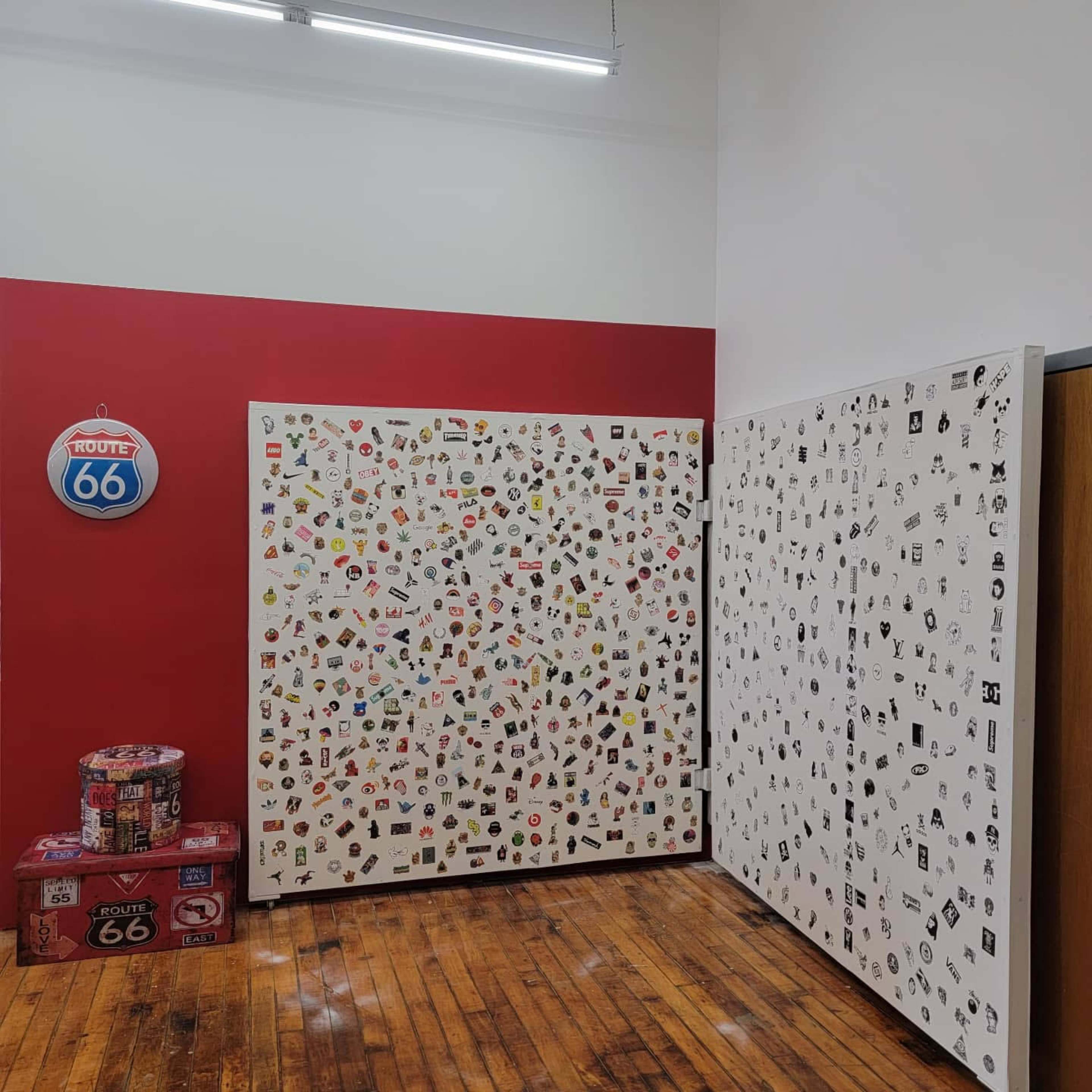 A room with red walls and white panels covered in various colorful stickers, alongside a circular Route 66 sign and stacked boxes featuring the Route 66 theme.
