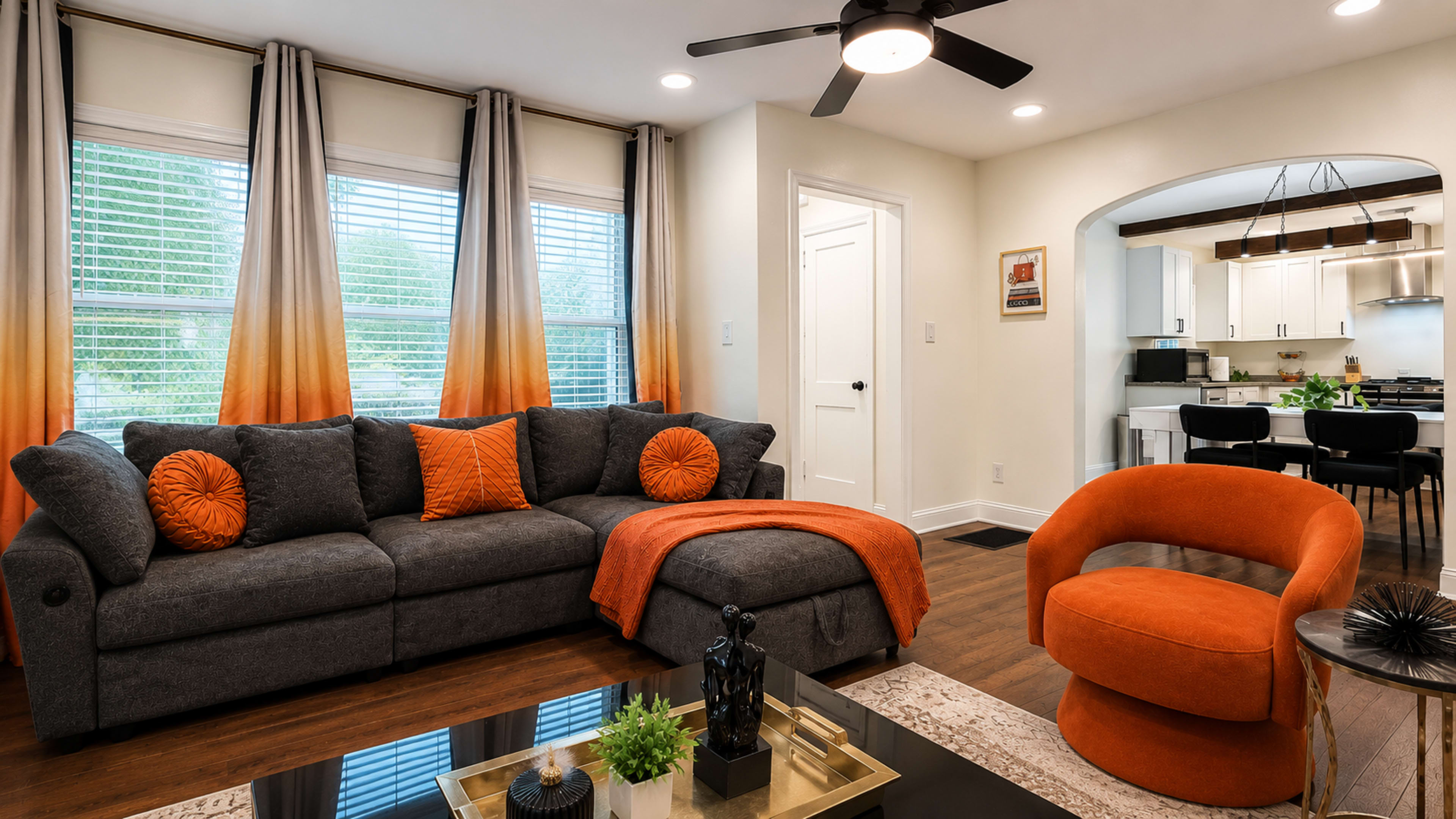 The image shows a modern living room featuring a dark sectional sofa with decorative orange pillows, an orange accent chair, and large windows covered with gradient curtains.