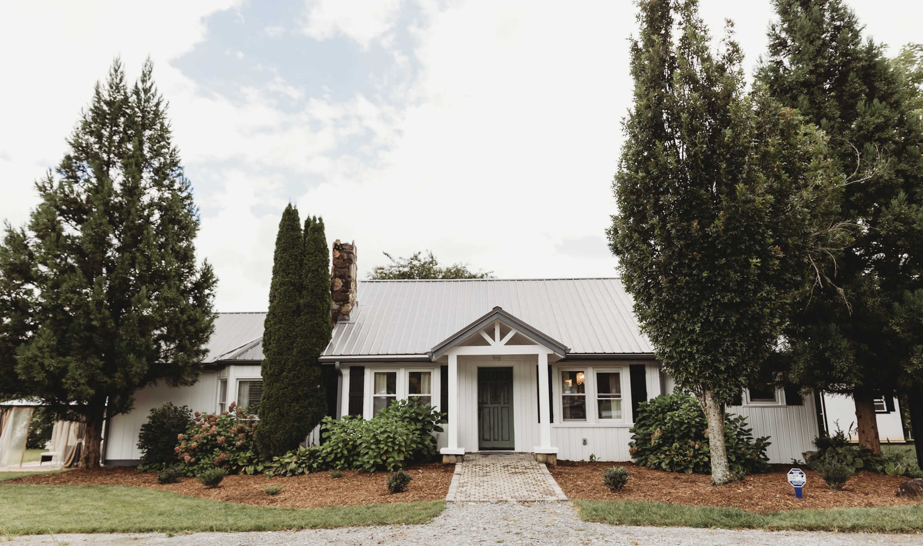 A single-story house with a gray metal roof, surrounded by trees and shrubs, features a central front door framed by two tall conifers.