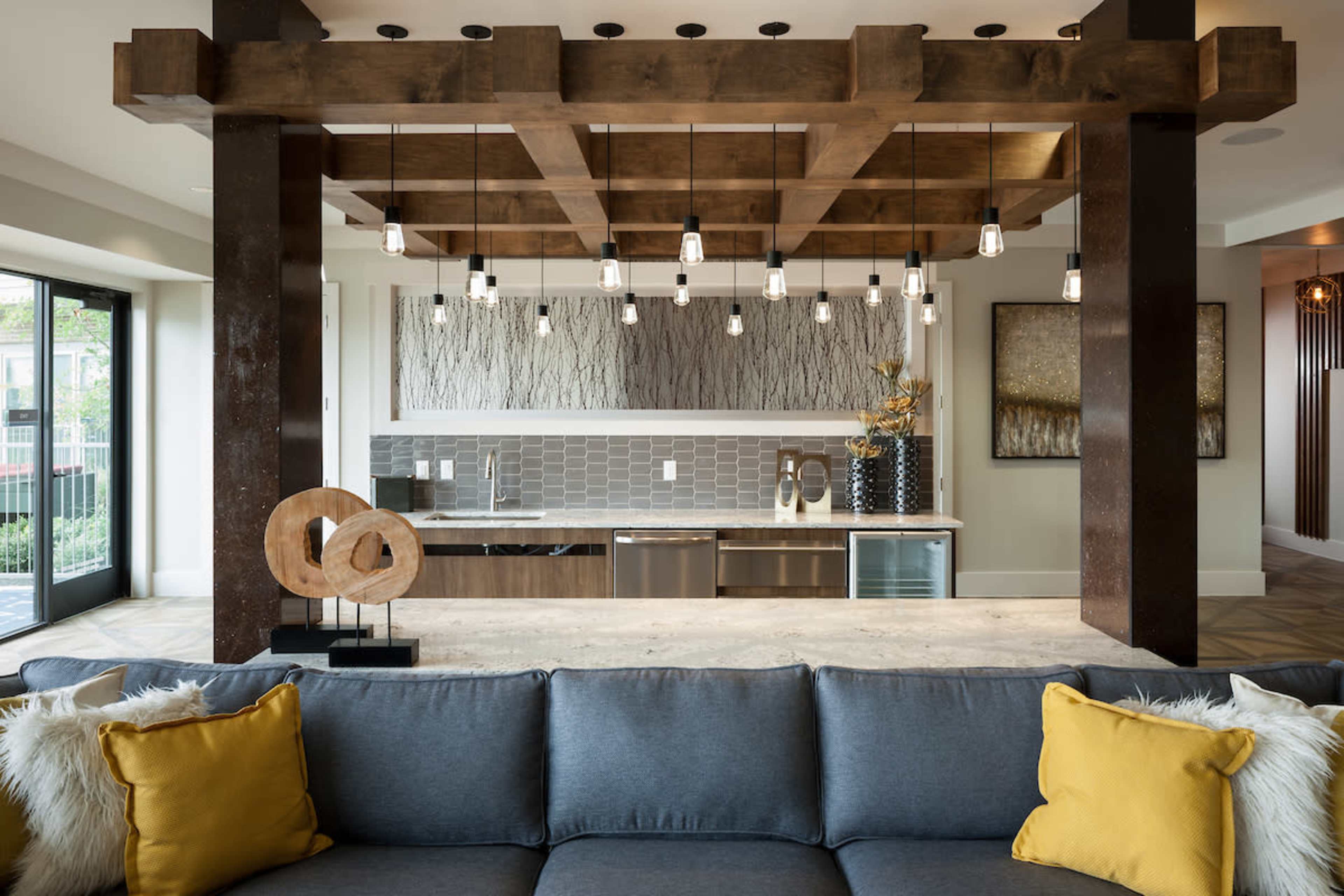 A modern interior space featuring a kitchen with a large wooden beam ceiling, pendant lights, and a cozy living area with a gray sofa adorned with yellow pillows.