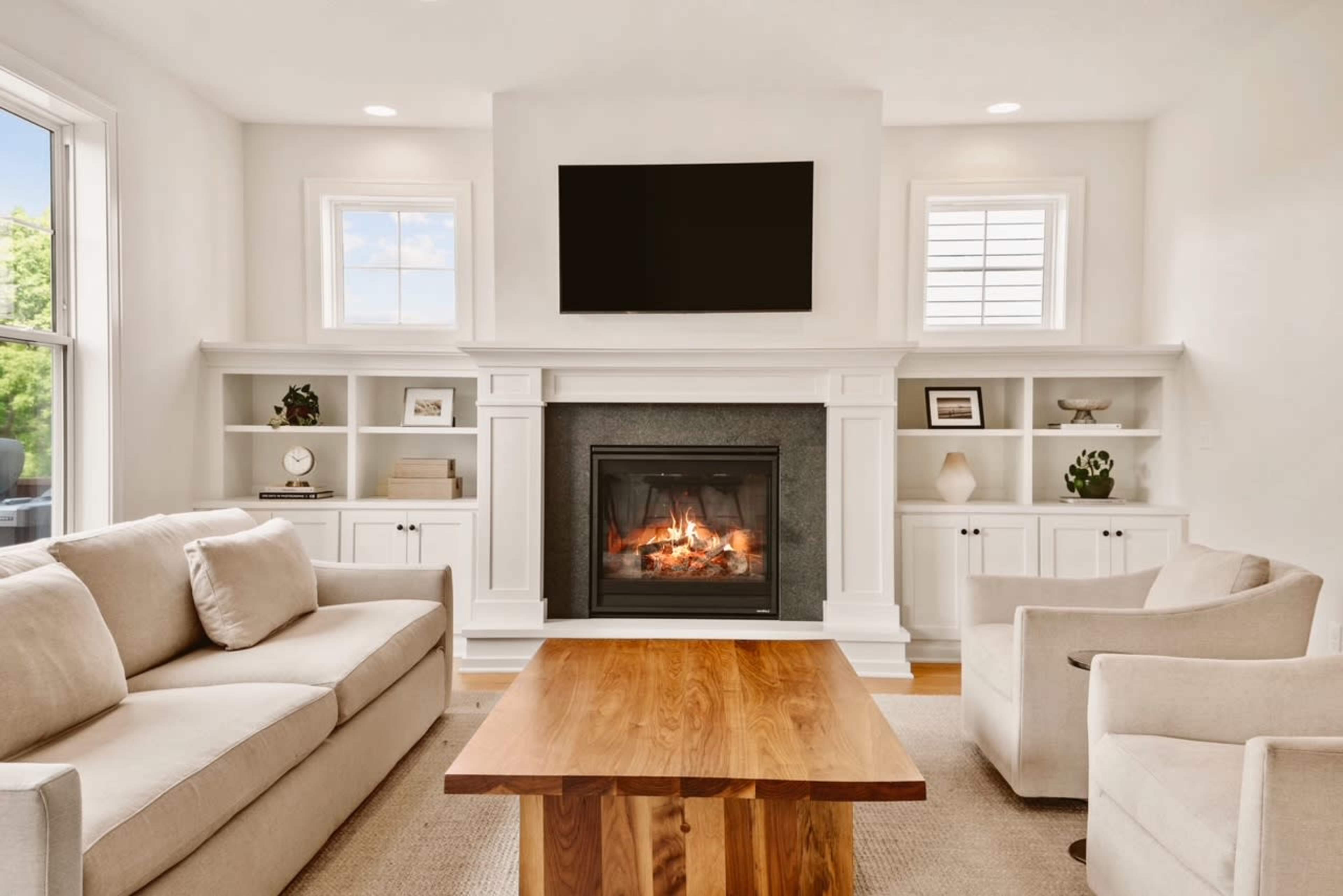 The image shows a modern living room with a beige sofa, two armchairs, a wooden coffee table, and a fireplace built into a white mantel, featuring a flat-screen TV above it.