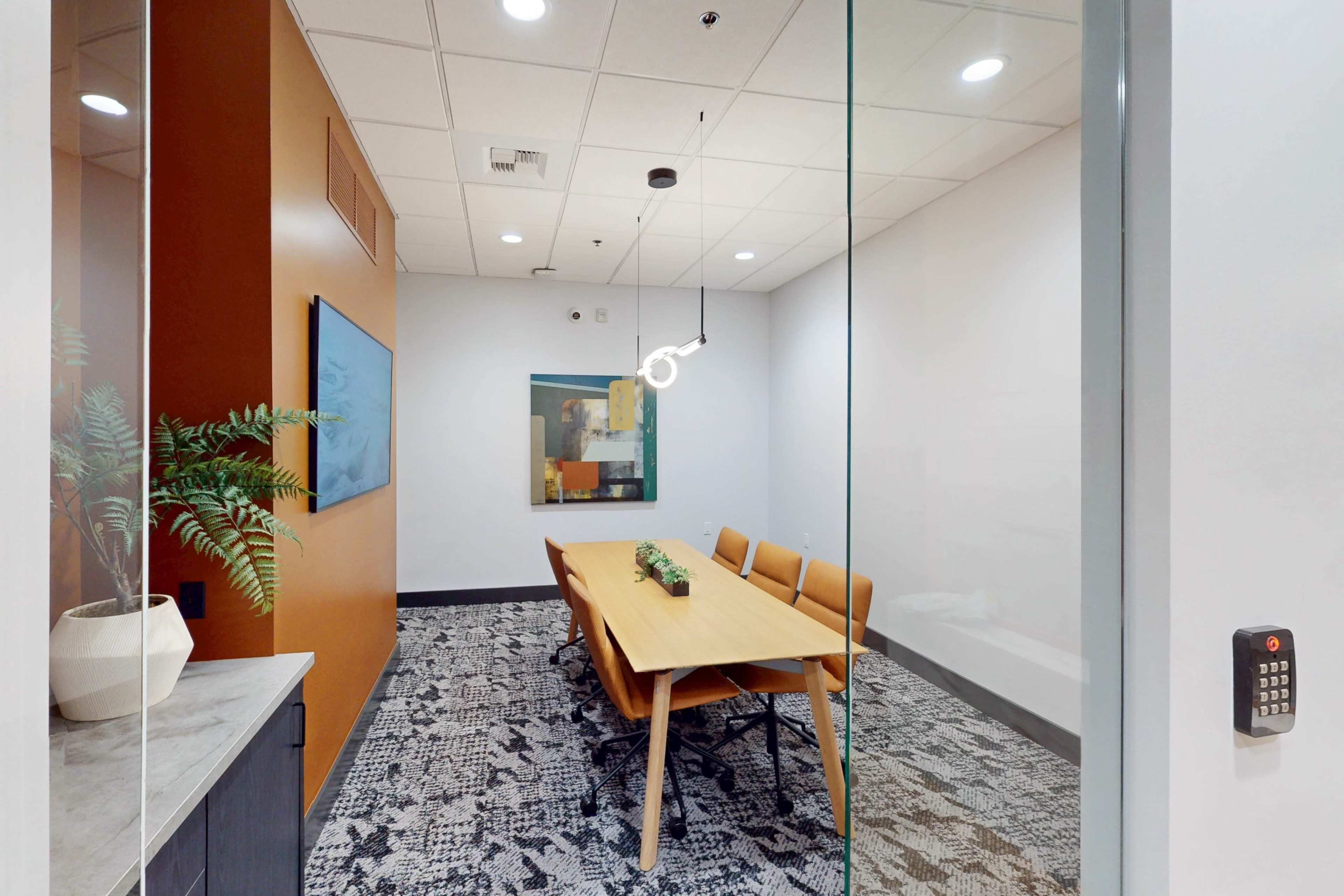 A modern conference room features a rectangular wooden table surrounded by four brown chairs and is partially enclosed by glass walls.