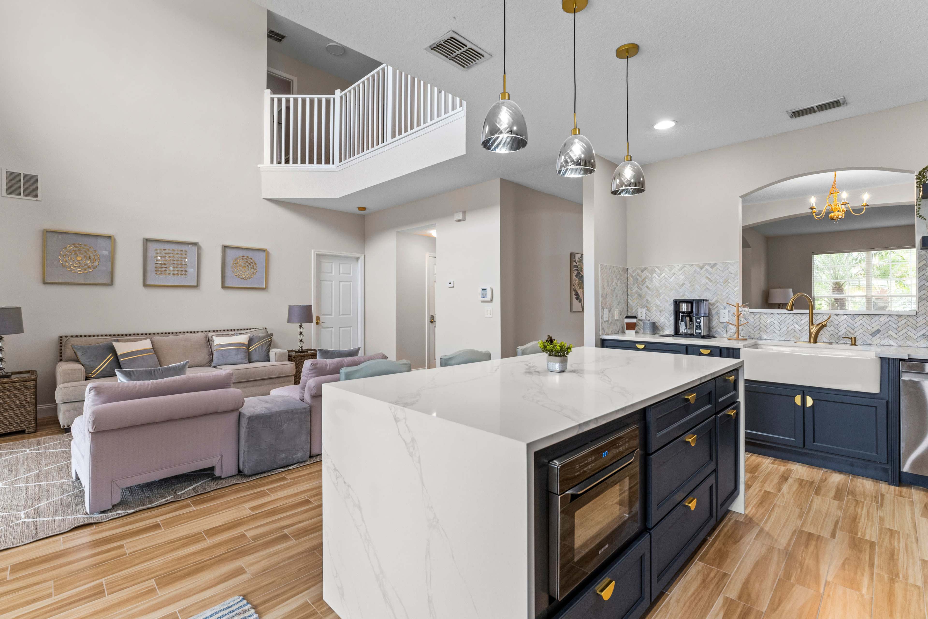 The image features a modern kitchen with a large island, pendant lighting, and a living area in the background, showcasing a light color scheme and comfortable seating.