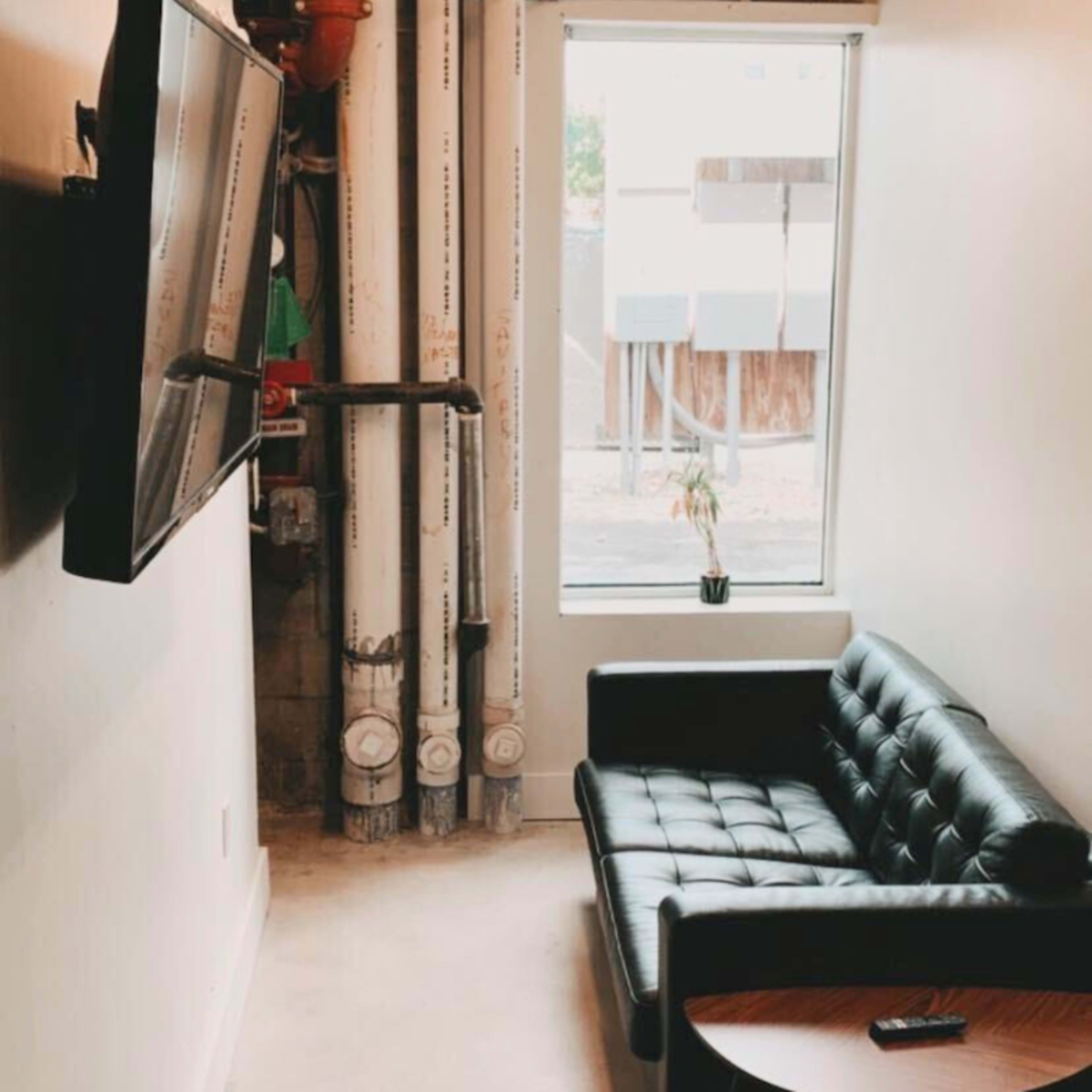 A black leather couch sits beside a window with a small plant, next to exposed pipes and a wall-mounted television.