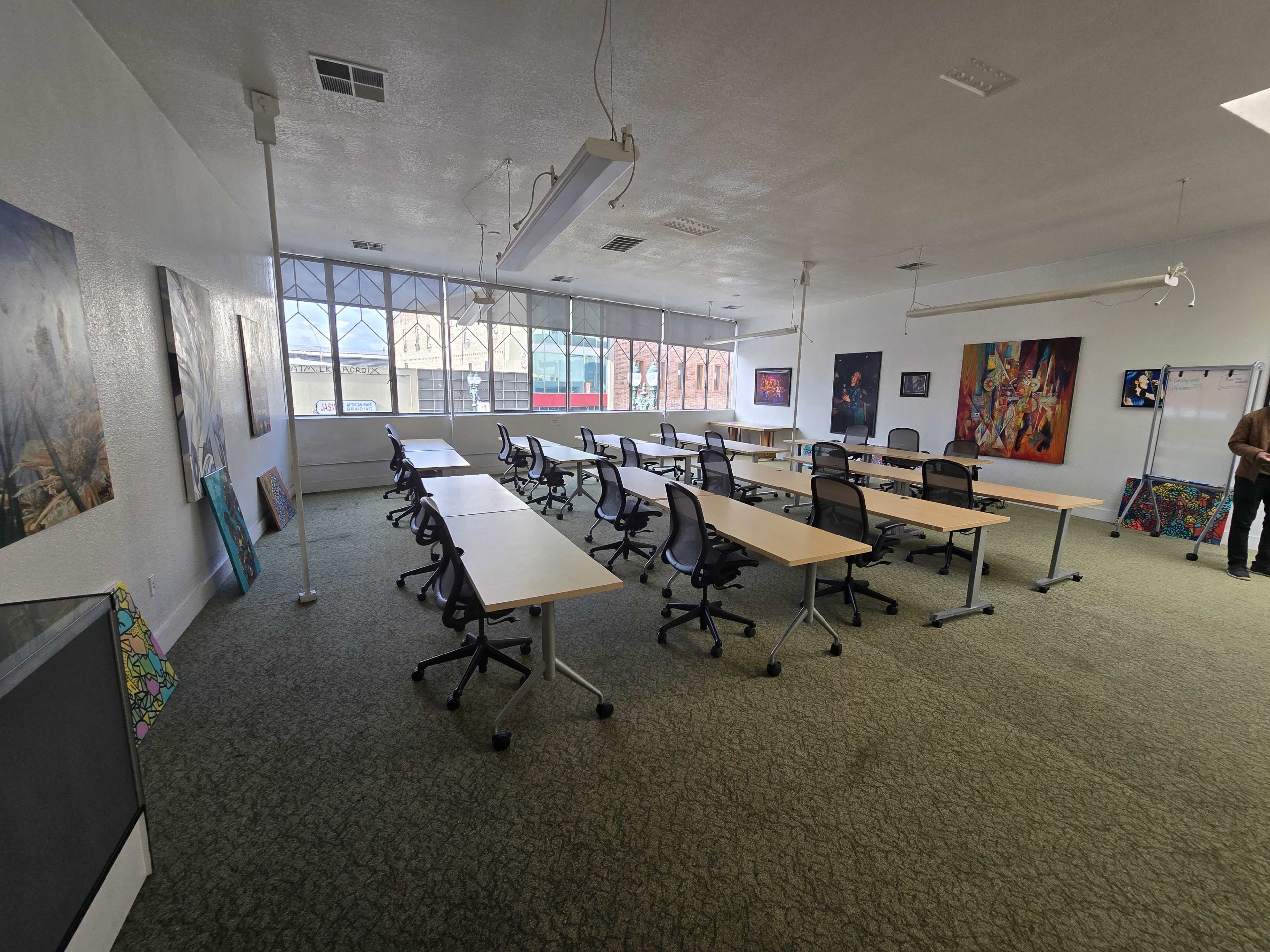 The image shows an empty conference room with multiple rectangular tables and office chairs arranged in a grid layout, large windows along one wall, and colorful artwork displayed on the walls.