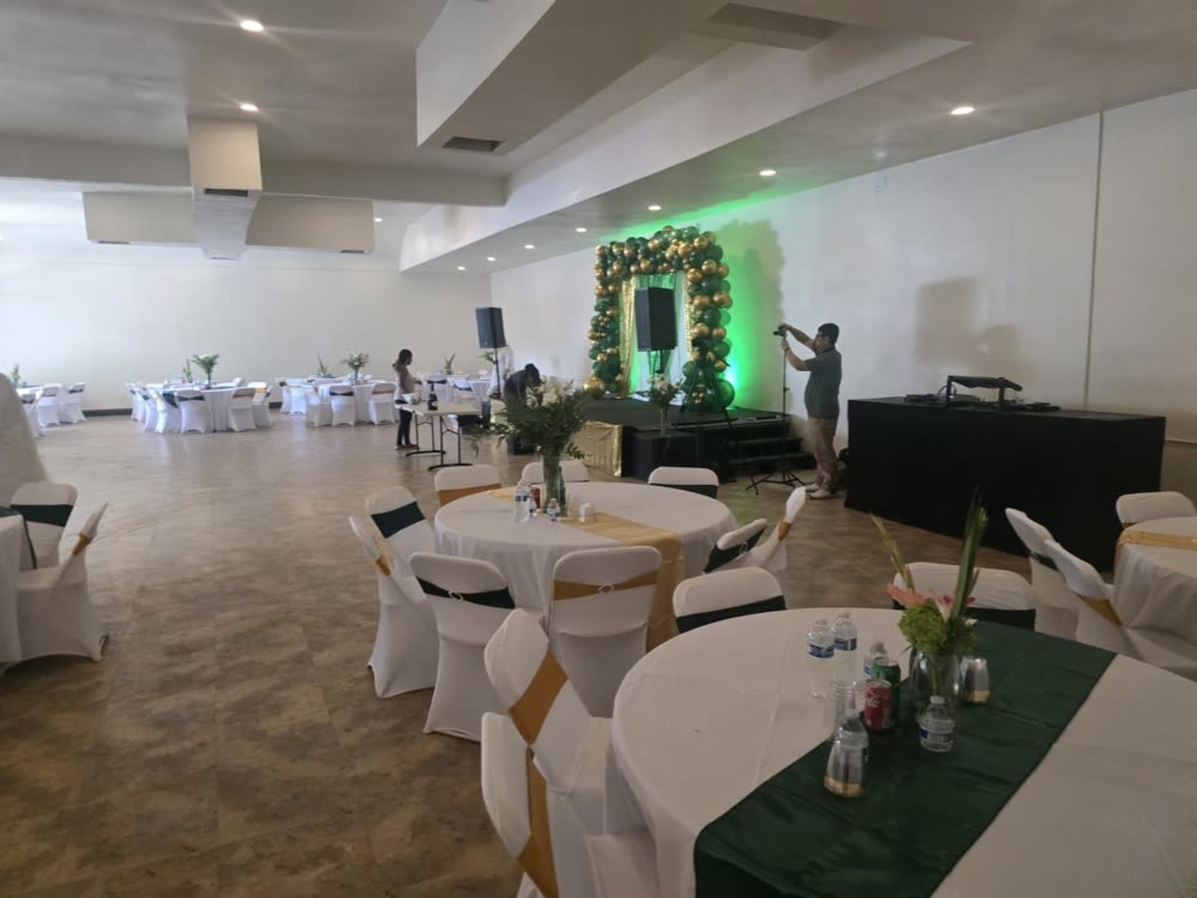 A spacious event hall is set up with round tables dressed in white and green, featuring floral centerpieces, and a stage adorned with green and gold decorations.