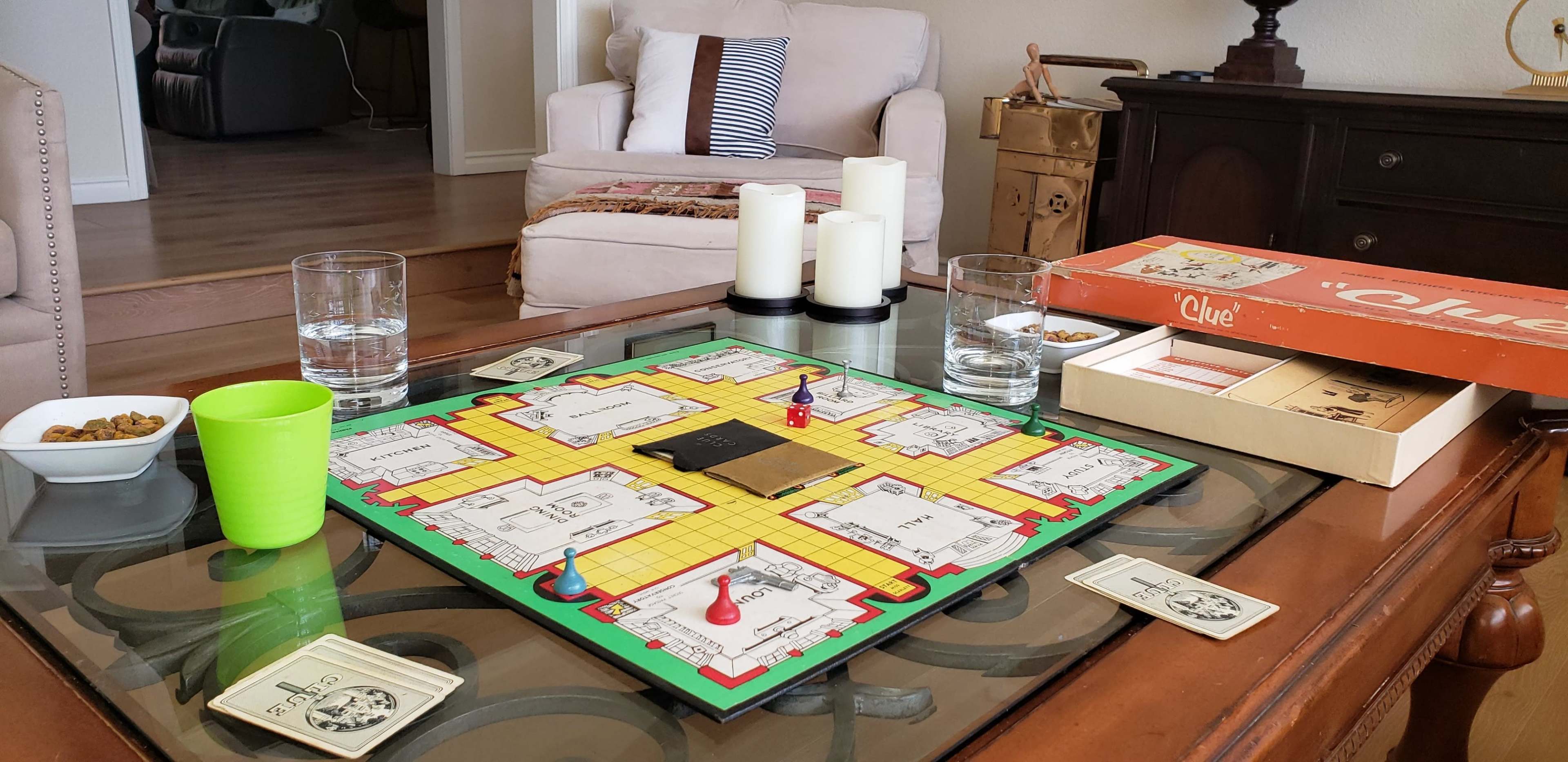 A Clue board game is set up on a table, accompanied by glasses of water, a small green cup, snacks, and lit candles.