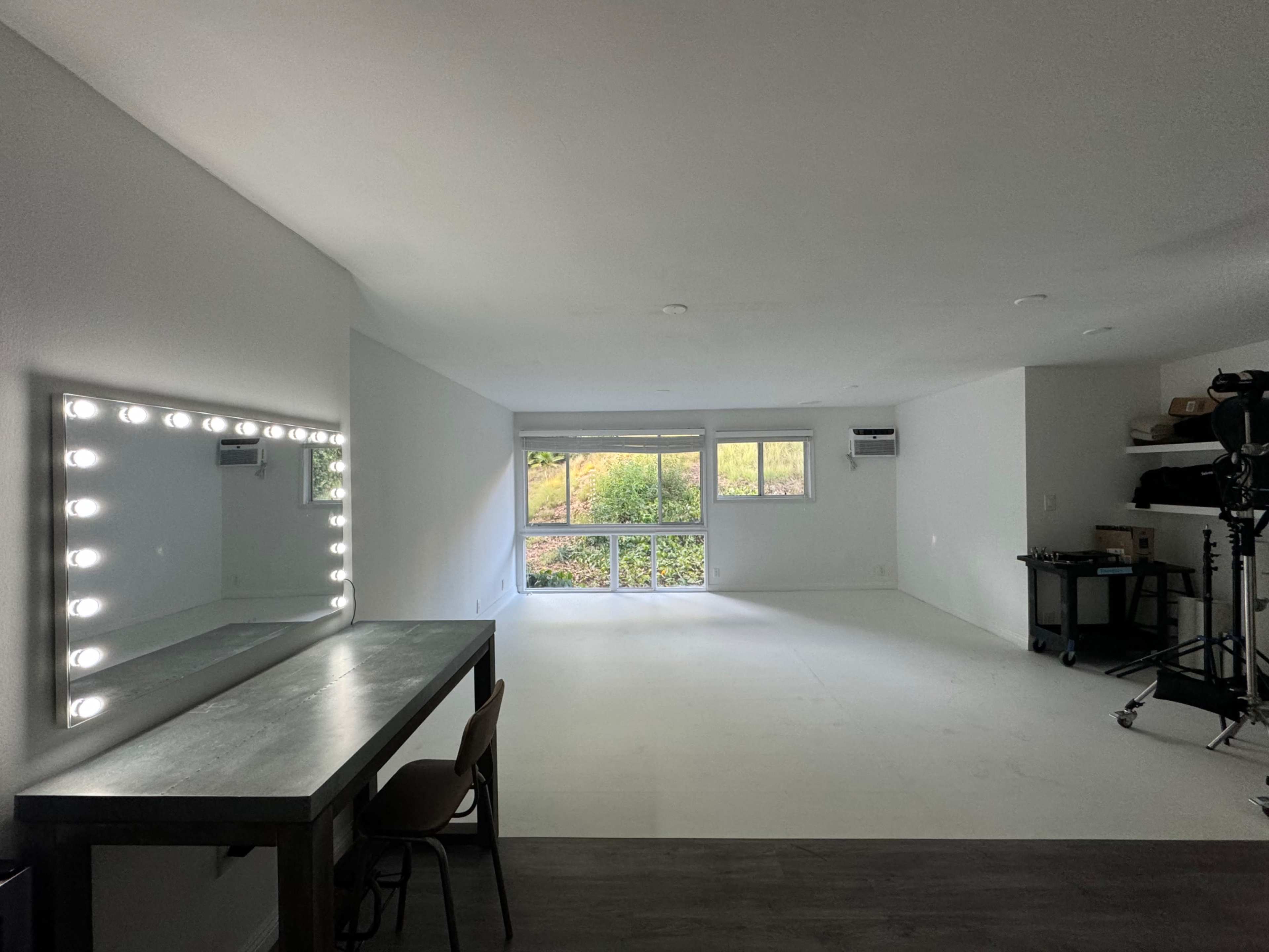 The image shows a spacious, empty room featuring a large mirror with light bulbs, a simple table with a chair, and a window overlooking a garden.