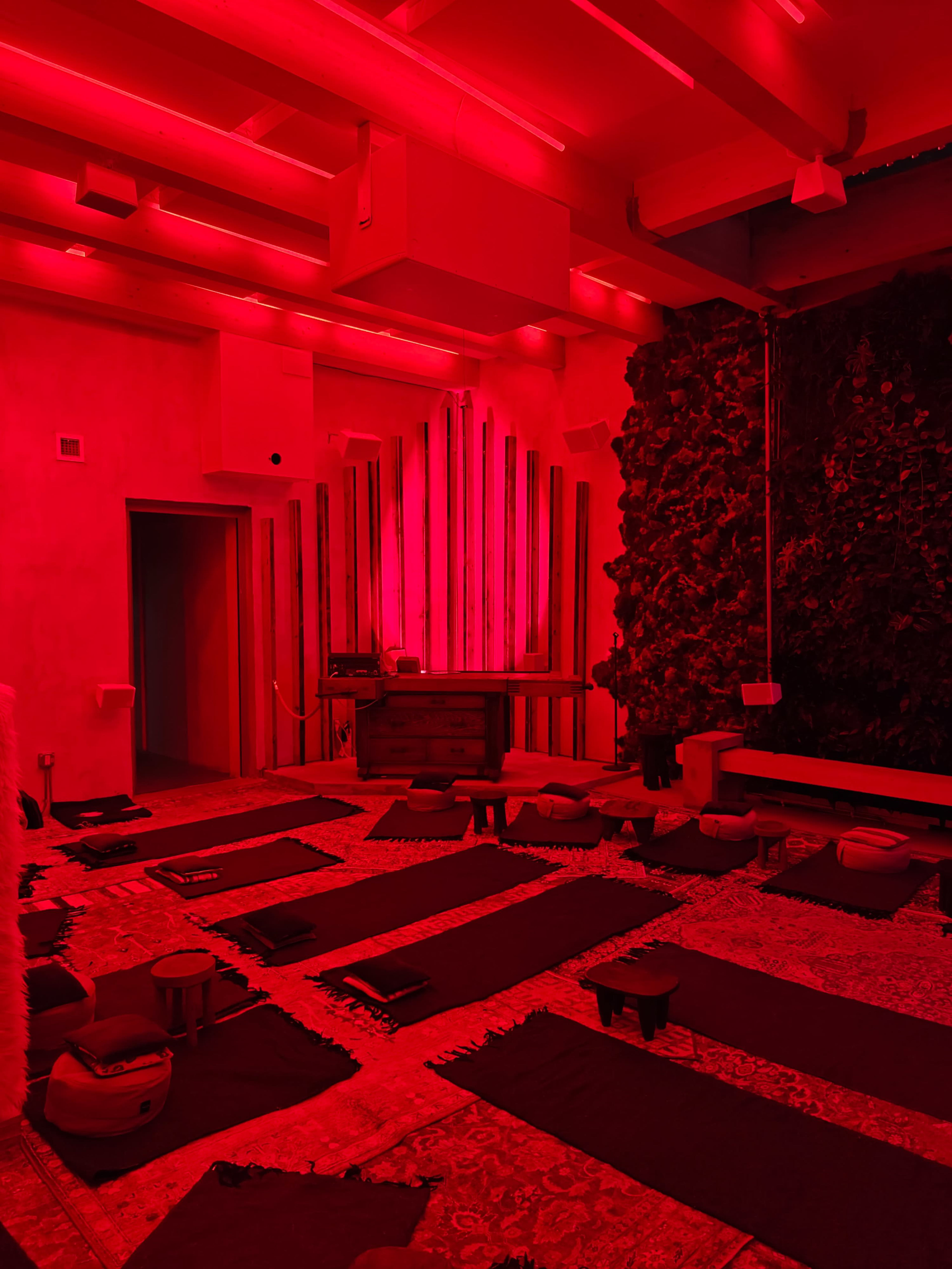 The room features red lighting, a wall covered in greenery, and several meditation mats arranged on the floor.