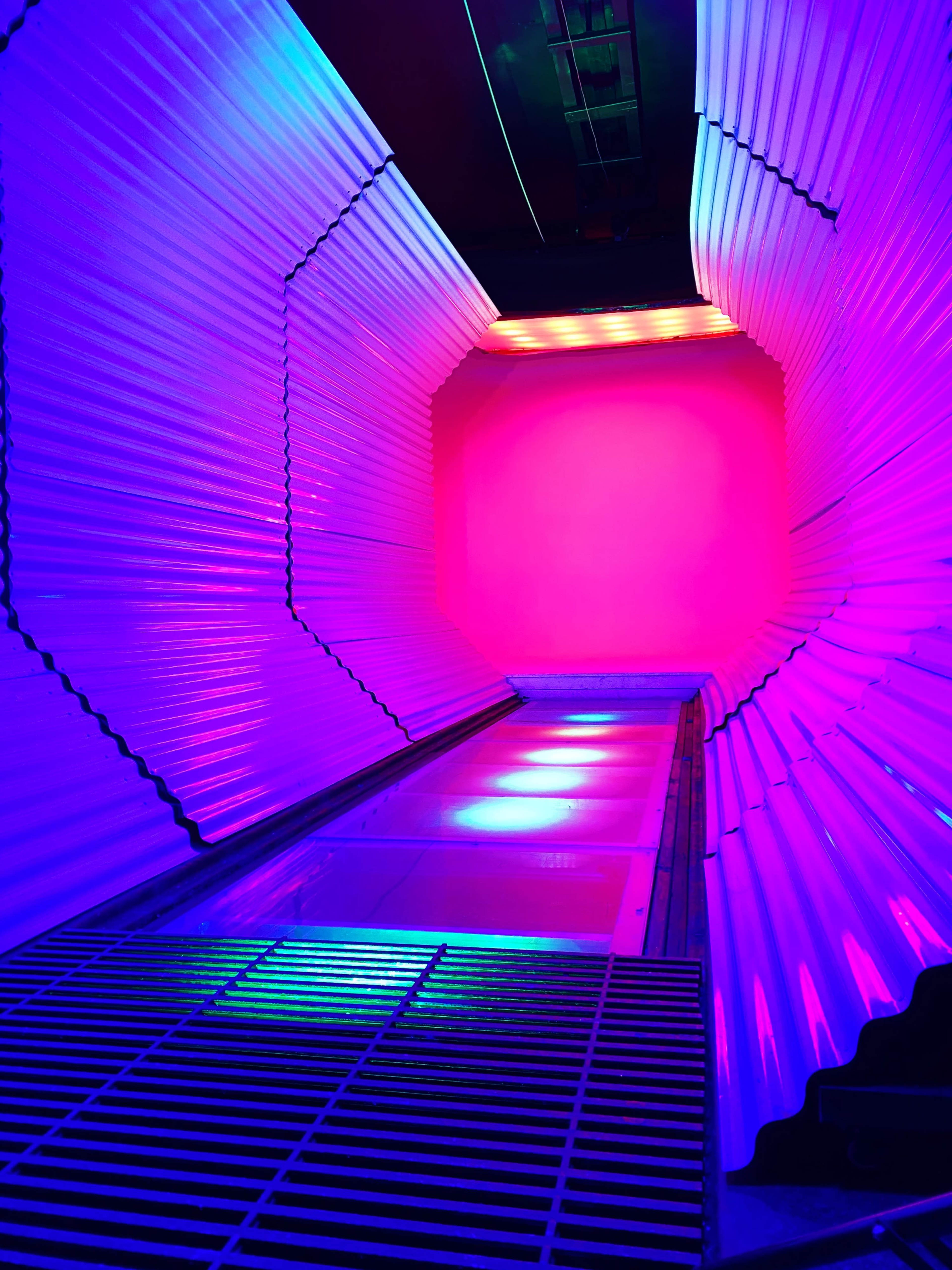 A brightly lit corridor with curved metallic walls and a vibrant pink and blue color scheme, leading to a solid wall at the end.