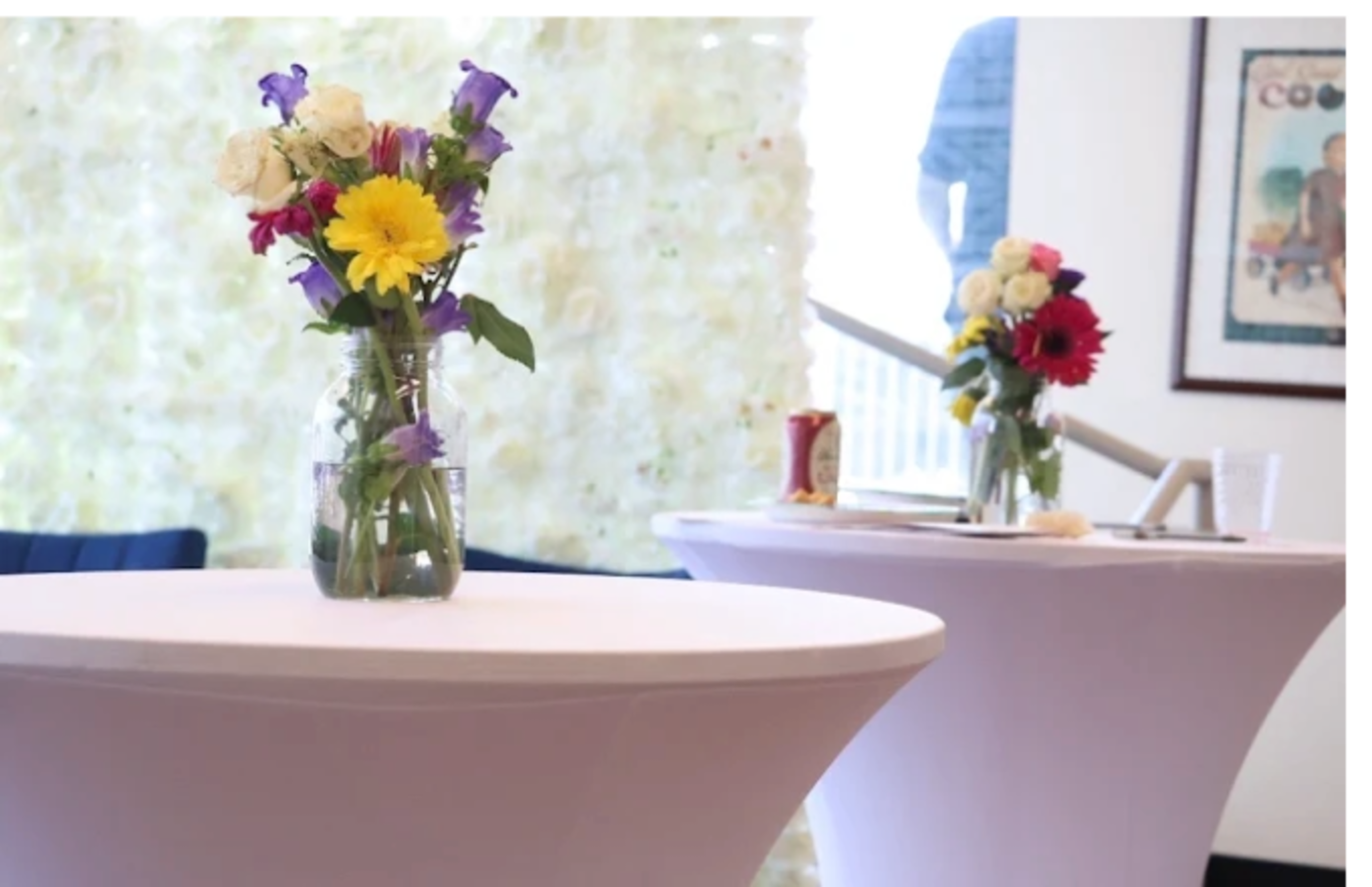 The image shows two round tables with floral arrangements in glass vases, set against a backdrop of a white floral wall.