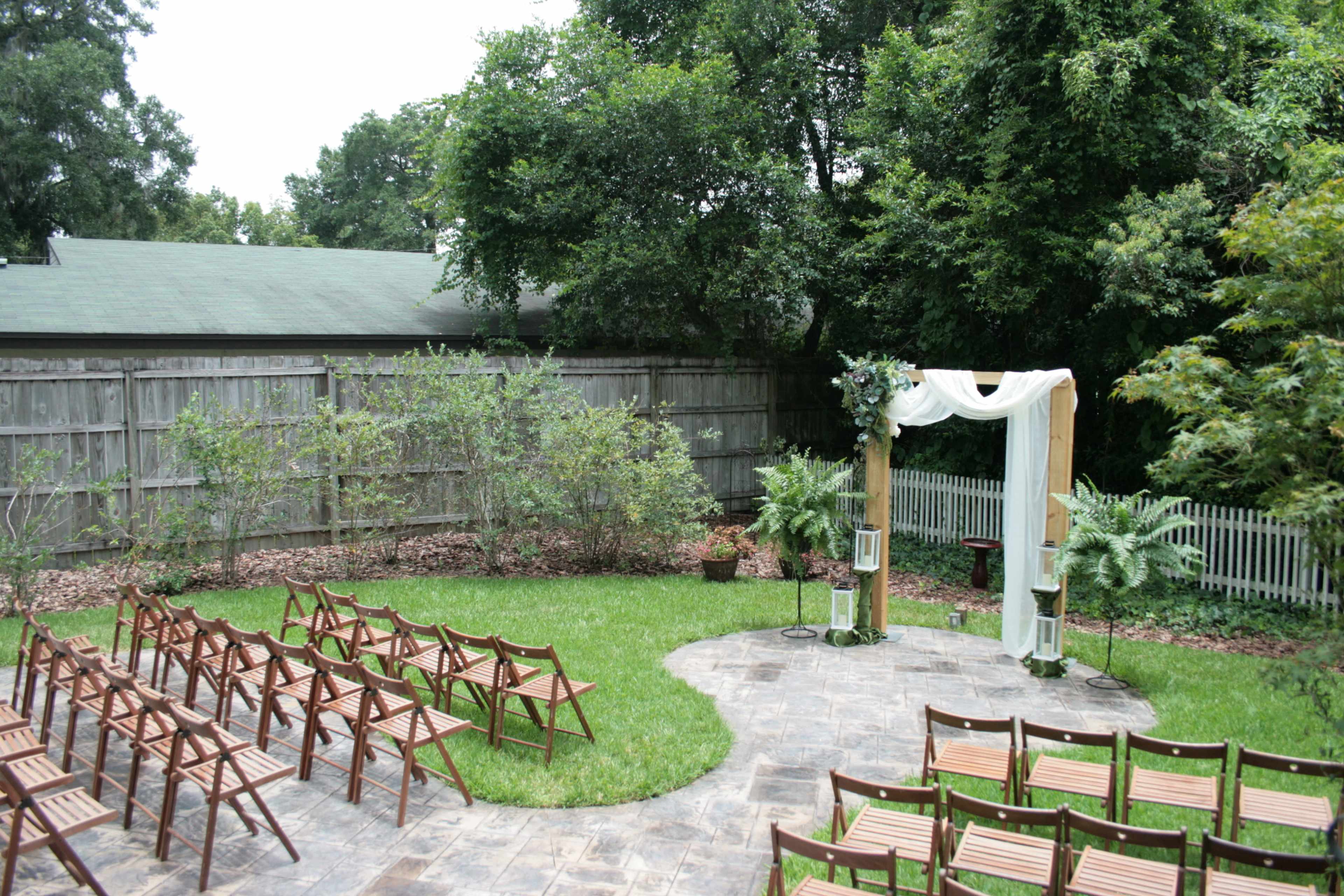 A wedding ceremony setup features wooden chairs arranged in rows facing a decorated archway surrounded by greenery in a garden area.