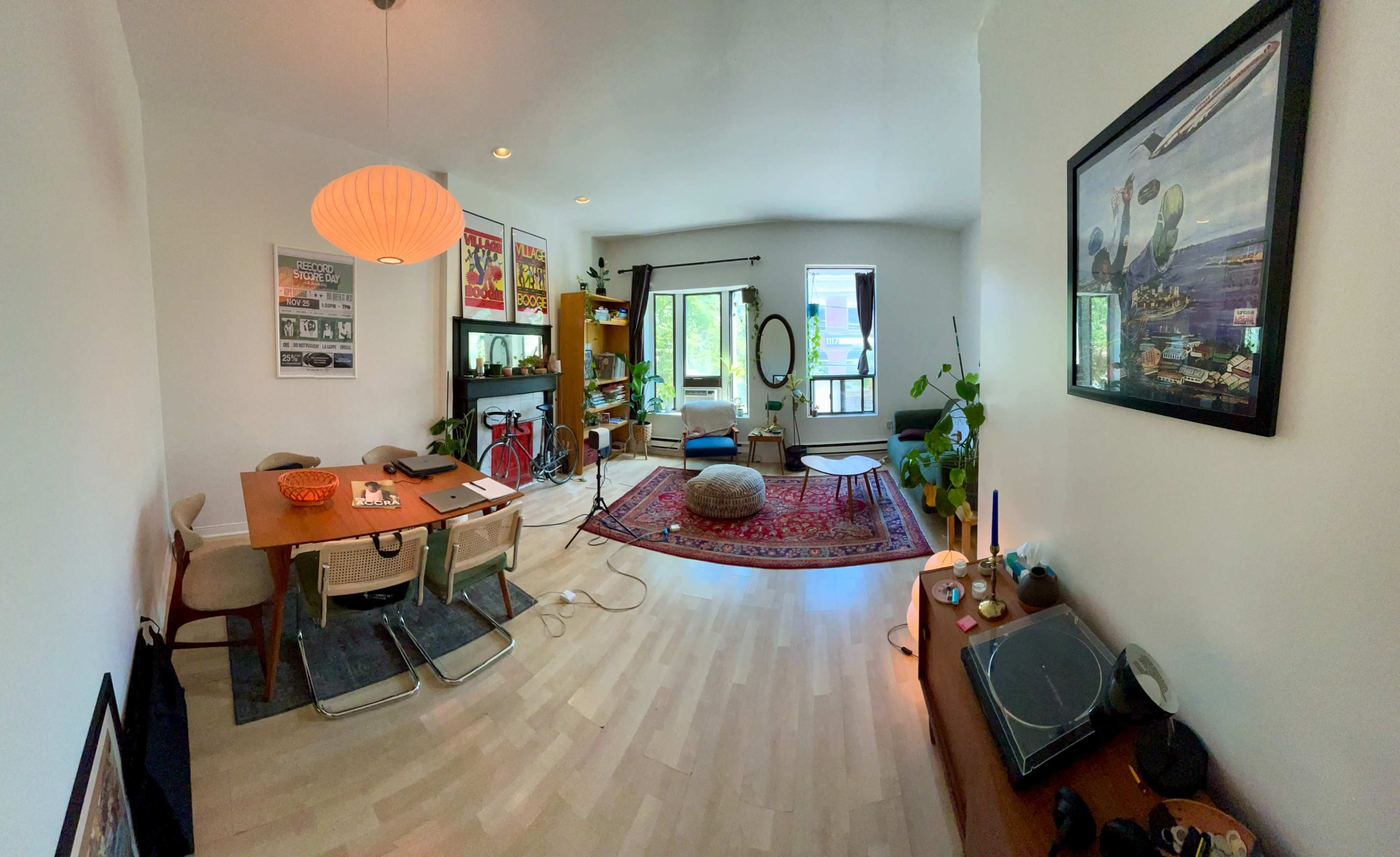 The image shows a well-lit living room with a dining area, featuring a round rug, a wooden table with chairs, plants, and artwork on the walls.
