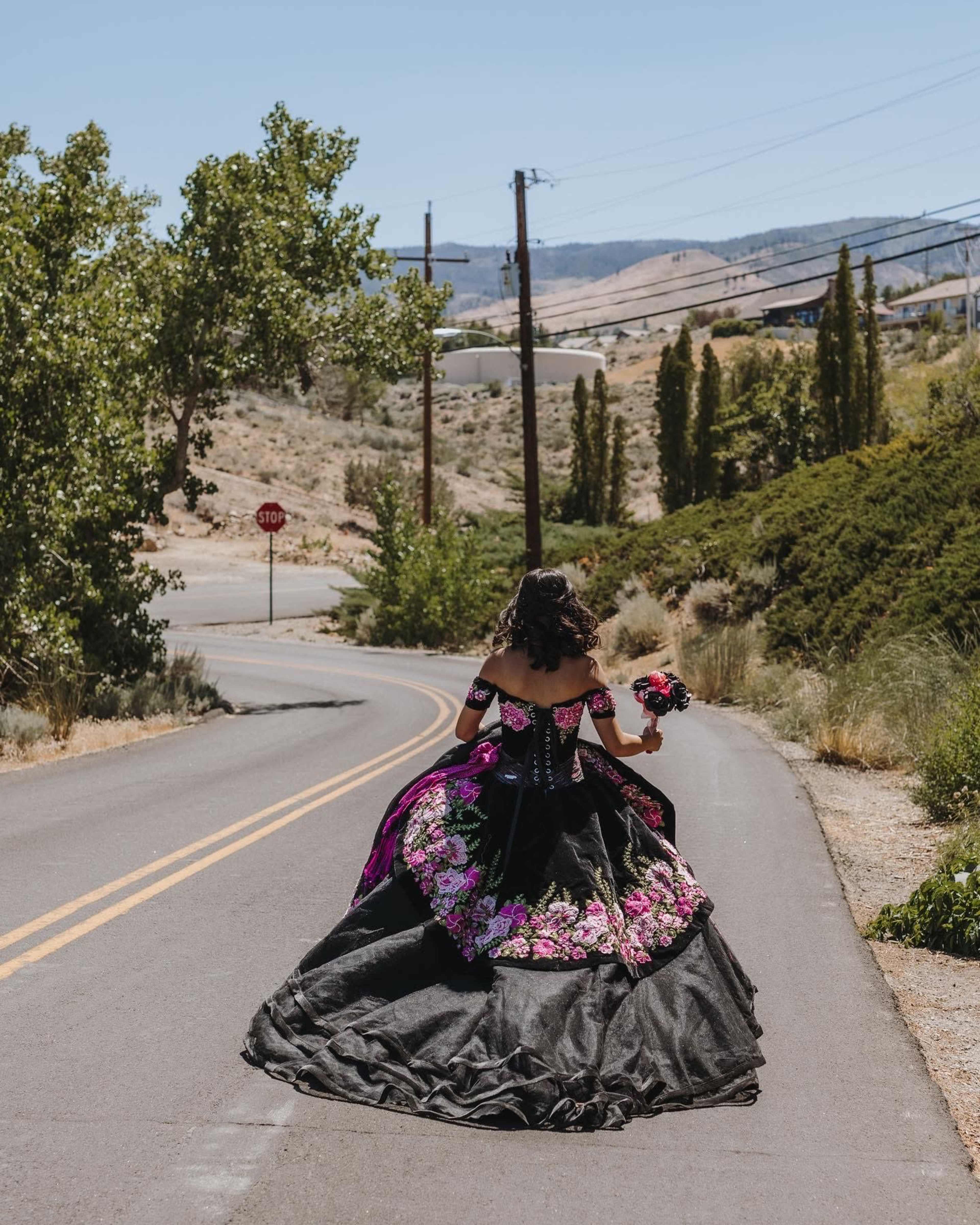 A woman in a black and pink floral gown walks along a rural road, holding a bouquet, with trees and hills in the background.