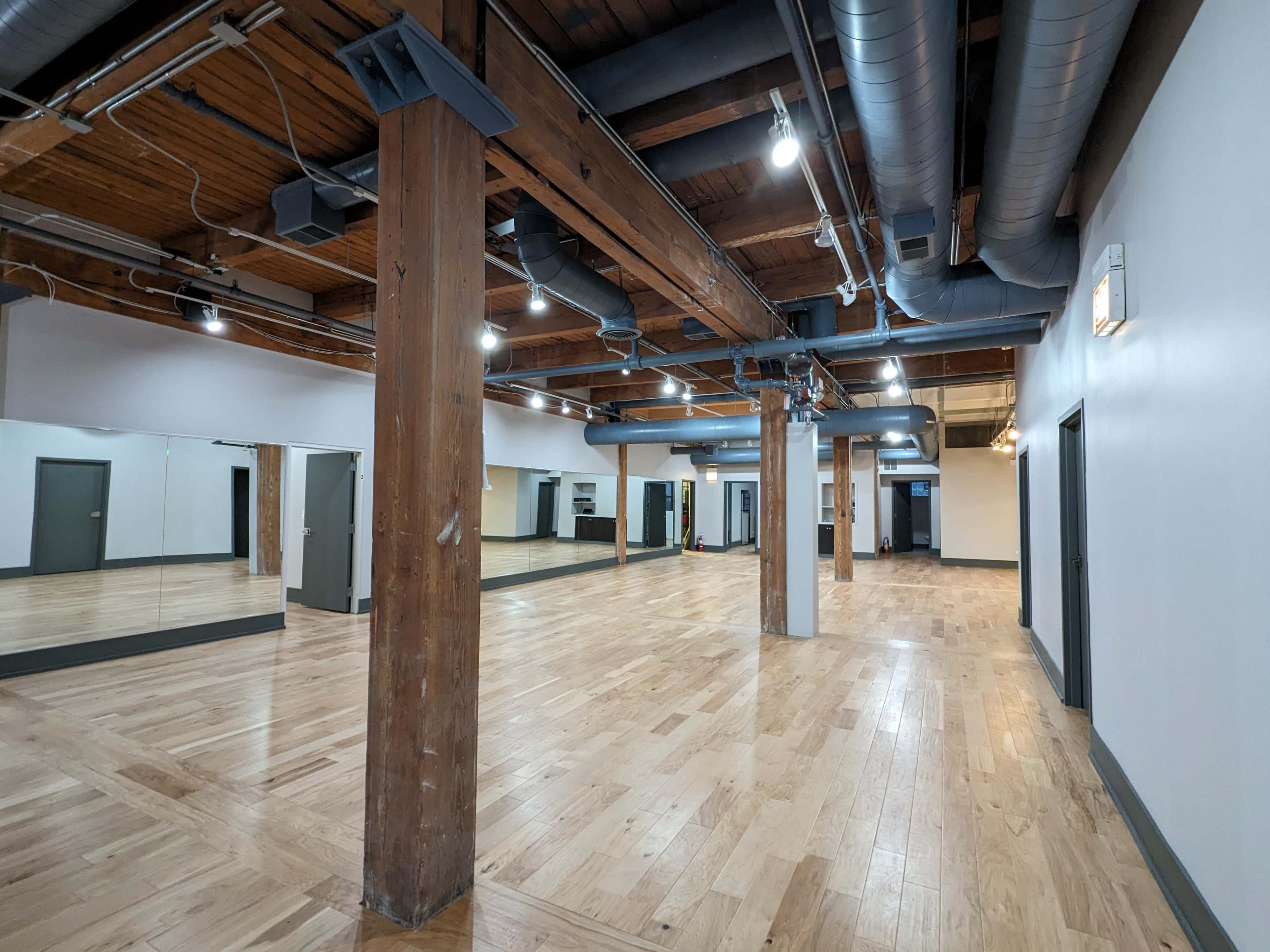 A spacious, empty wooden-floored studio with exposed overhead beams and large mirrors on one wall.