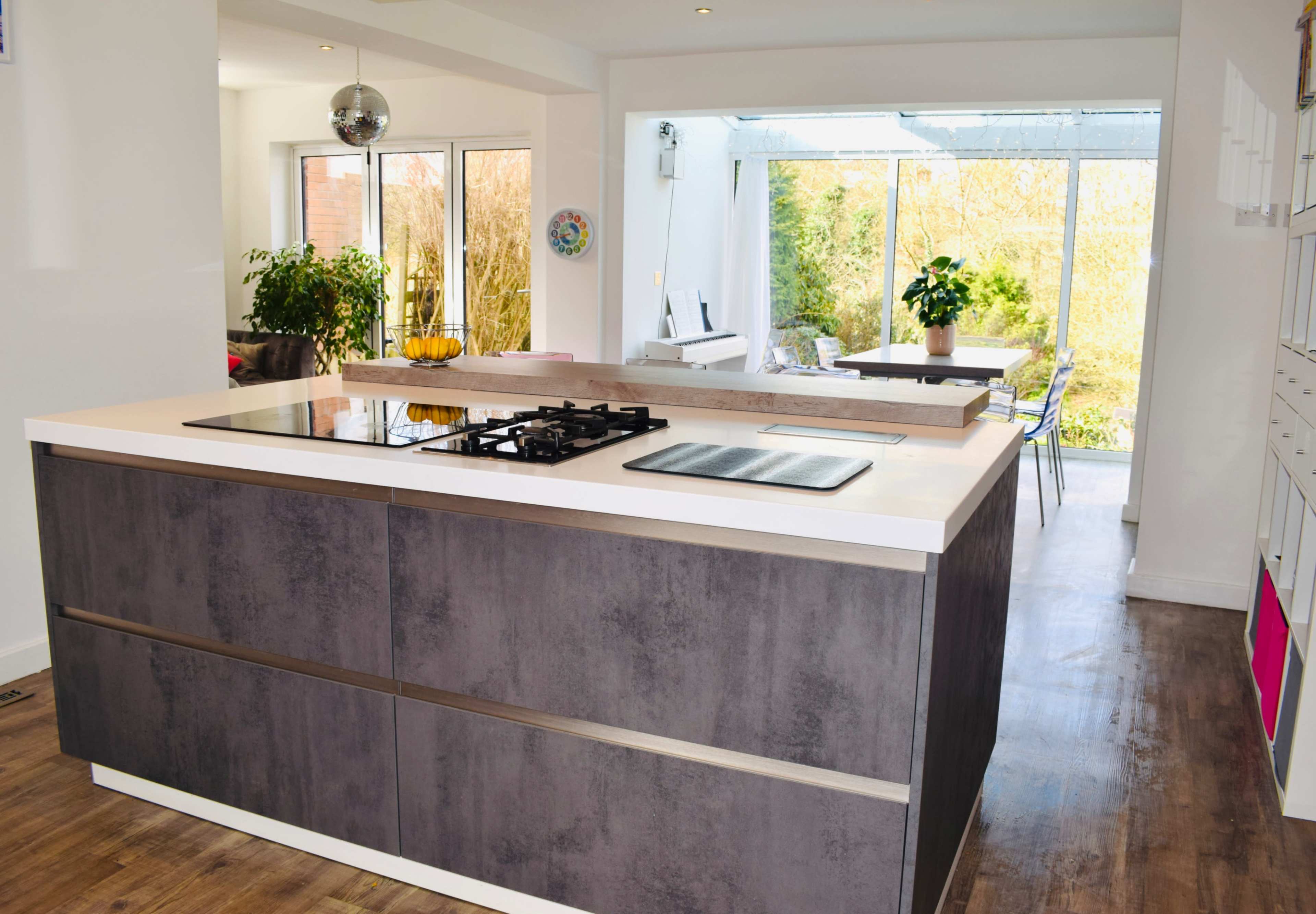 A modern kitchen with a central island featuring a built-in stovetop, surrounded by large windows that overlook a garden.