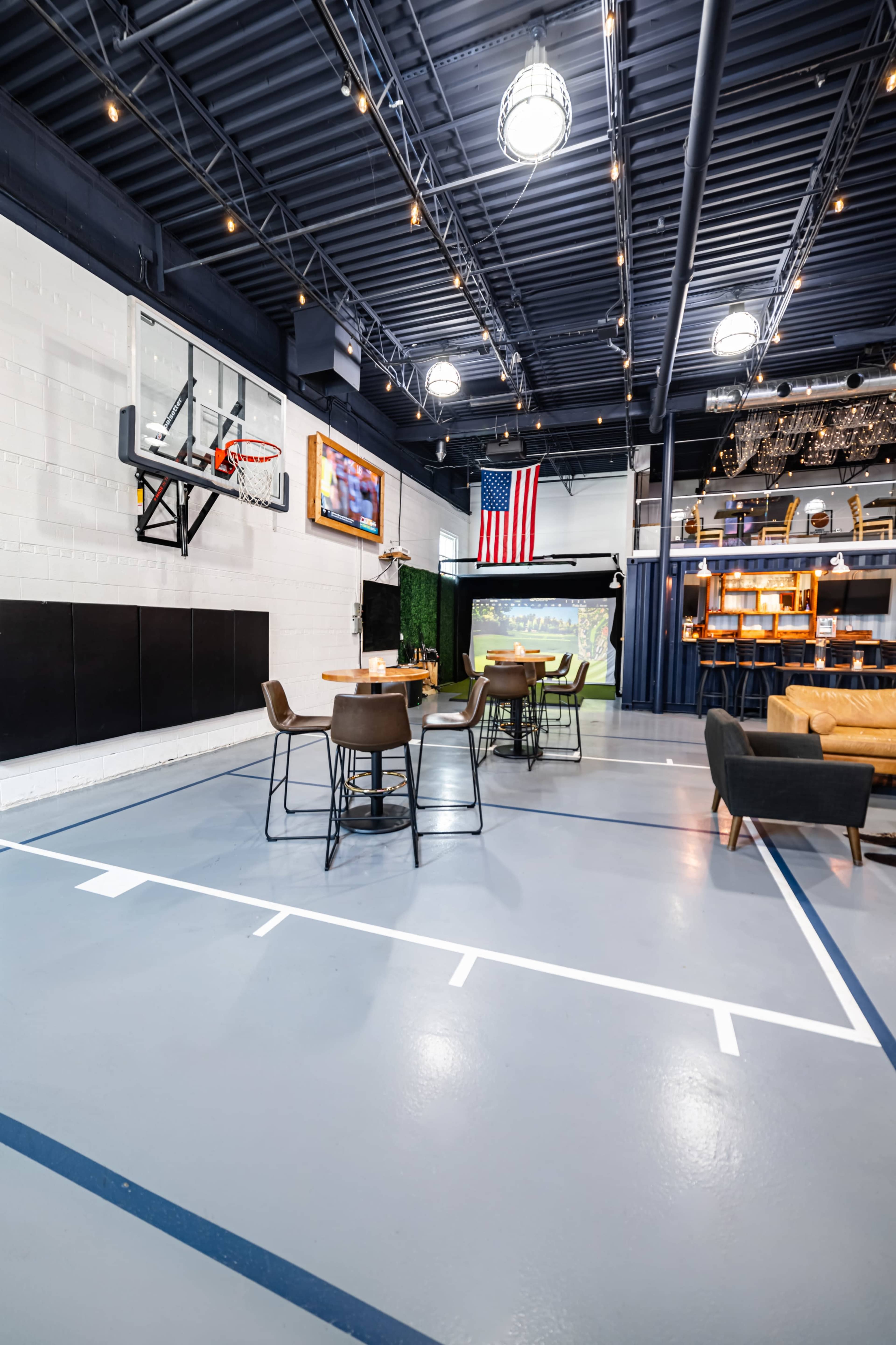 A spacious indoor area featuring a basketball hoop, a projector screen, several dining tables, and an American flag on the wall.