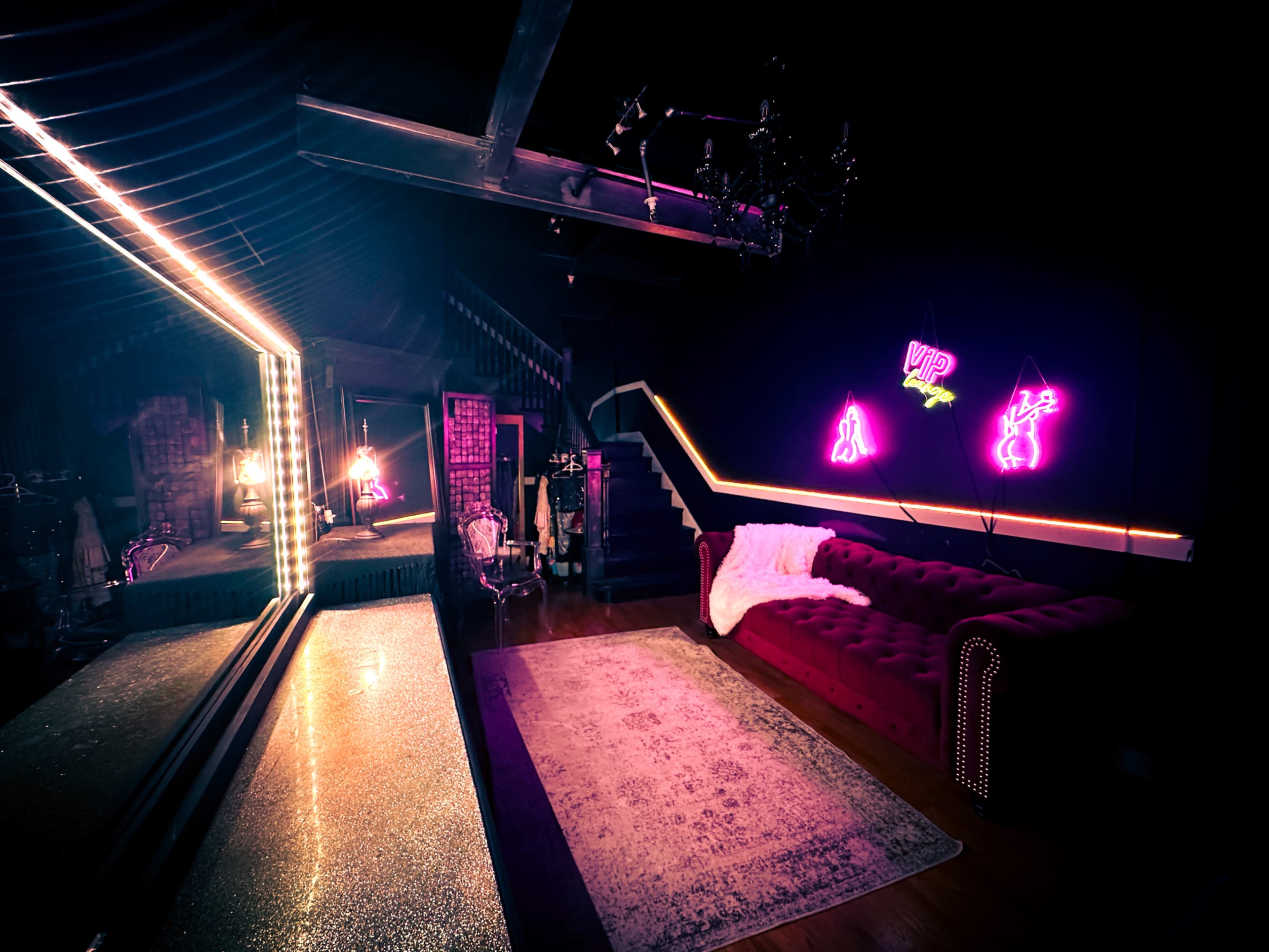 A dimly lit room with a plush red couch, a large mirror, and neon signs on the wall.