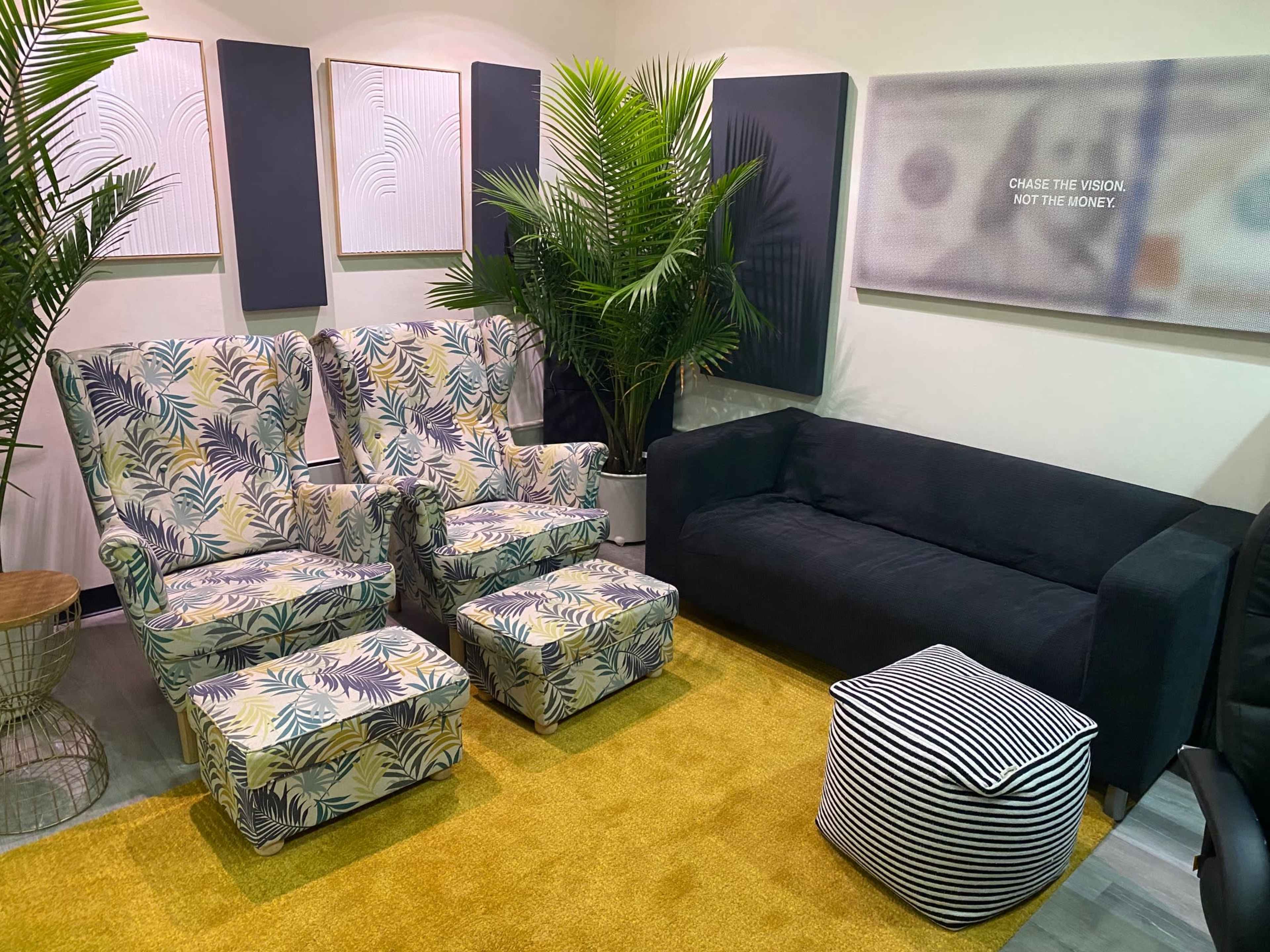 A cozy seating area featuring two patterned armchairs, a dark couch, a striped pouf, and tropical plants on a yellow rug.