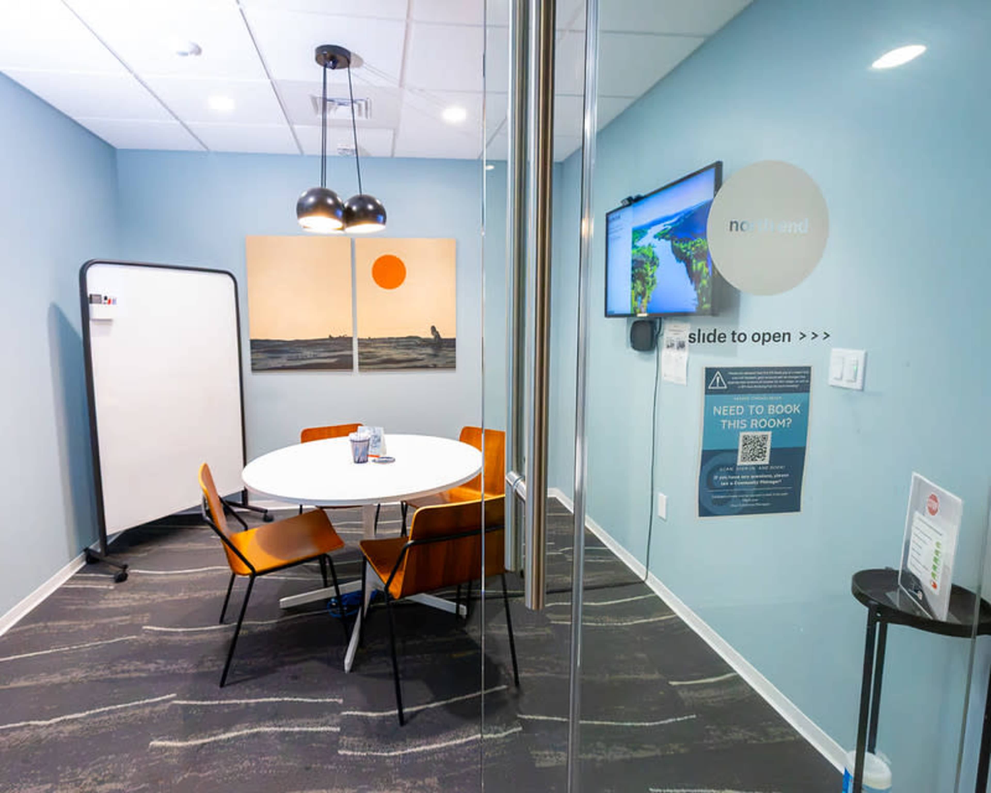 The image shows a small conference room with a round table, four chairs, and a wall-mounted TV, featuring artwork and a sliding glass door.