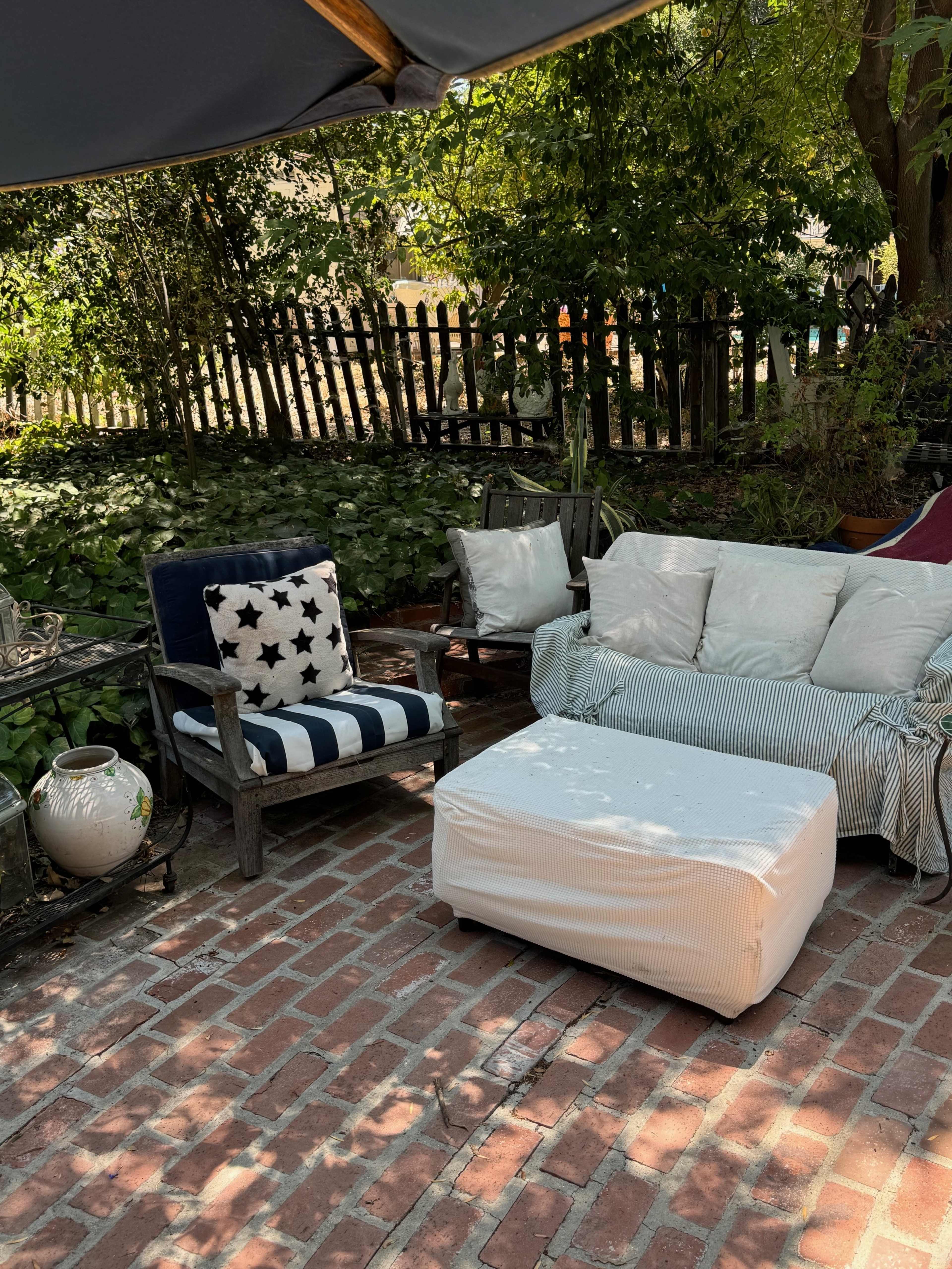 A small outdoor seating area features a striped couch, a star-patterned chair, and a rectangular ottoman on a brick patio surrounded by greenery.