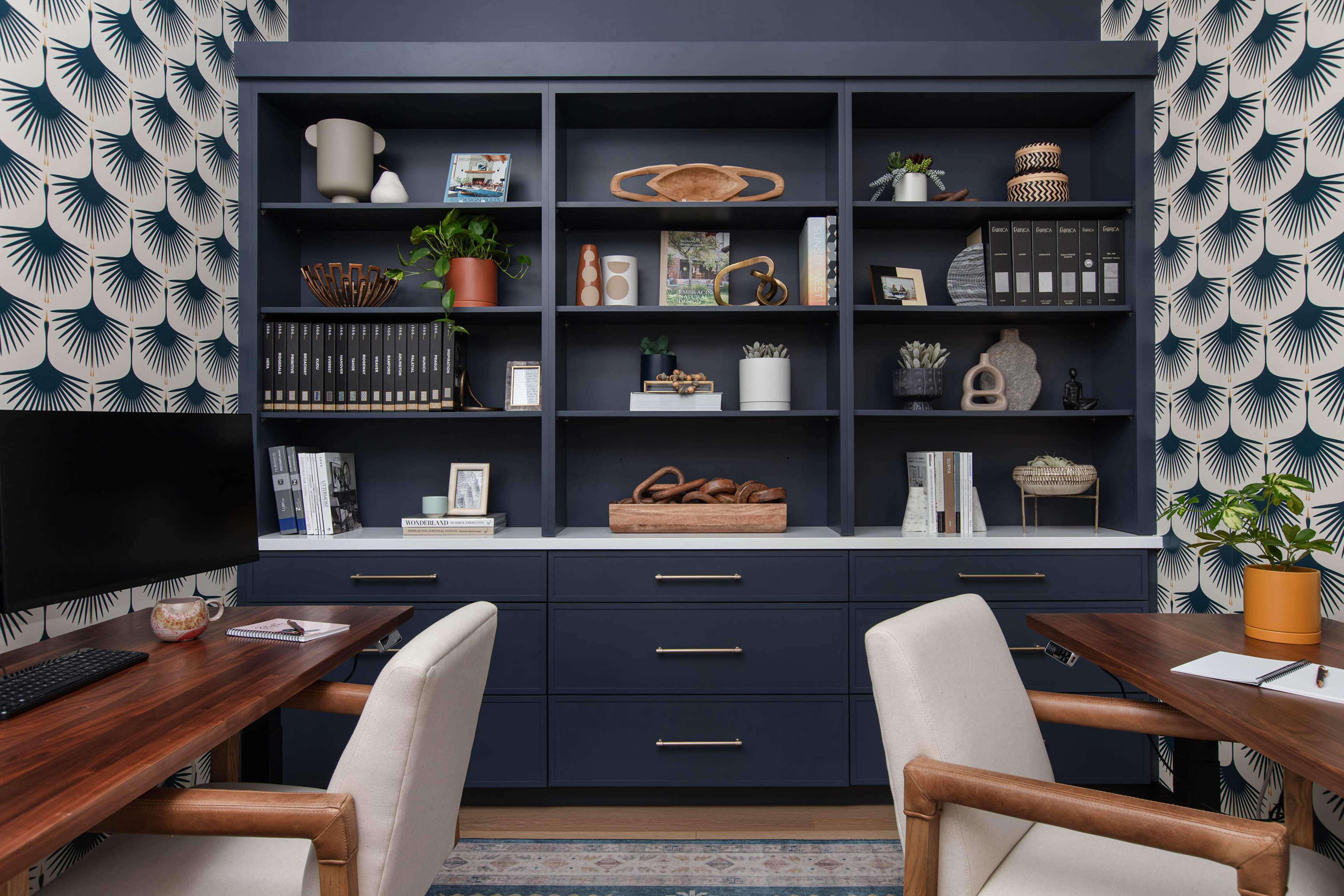 A modern office space featuring dark cabinetry with various decorative items and books, flanked by two wooden desks and a patterned wallpaper.
