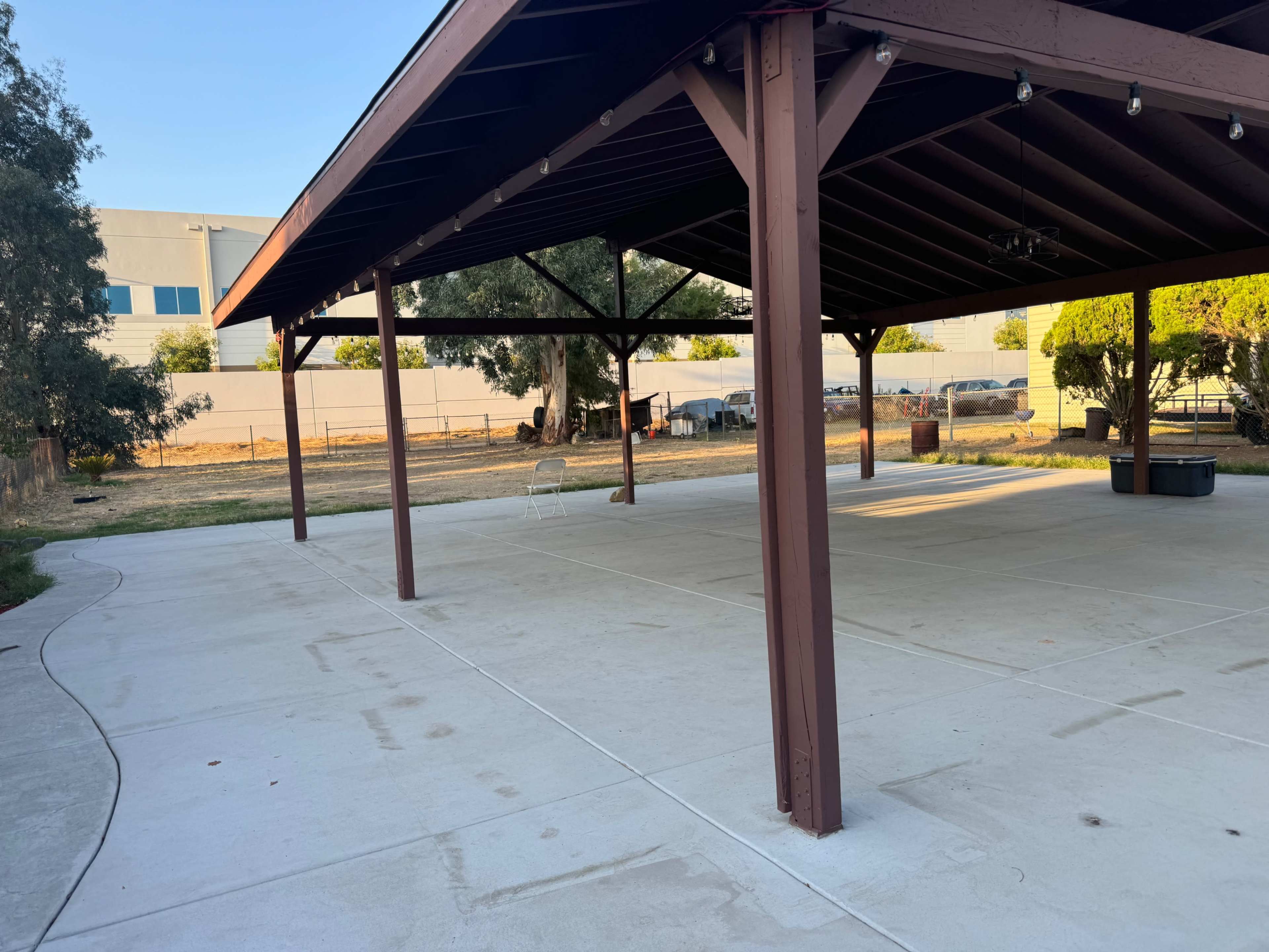 A large covered pavilion with wooden beams overlooks a grassy area and parked vehicles in the background.