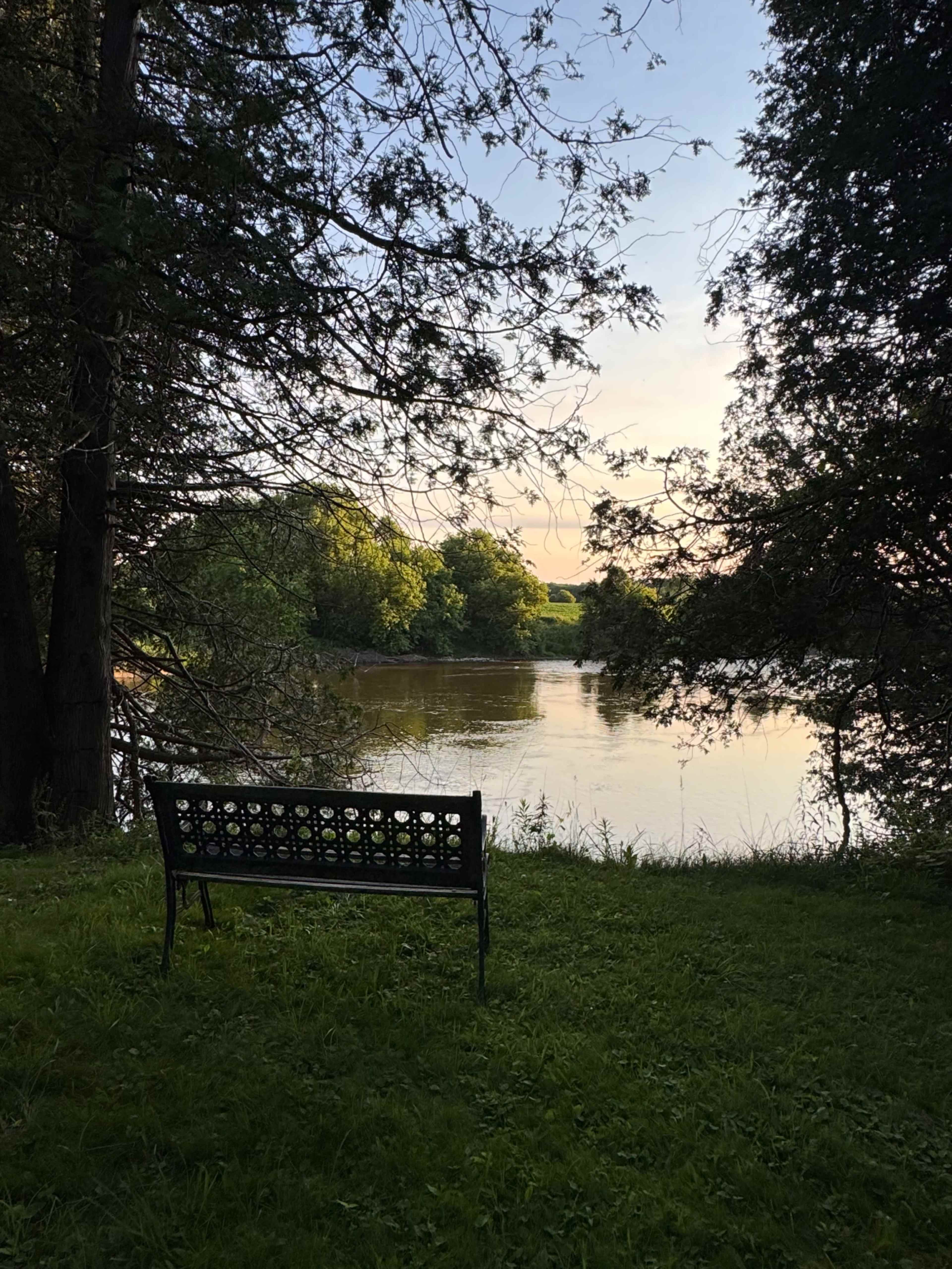 A metal bench is positioned near a quiet river, surrounded by trees and grass, with a soft sunset illuminating the scene.