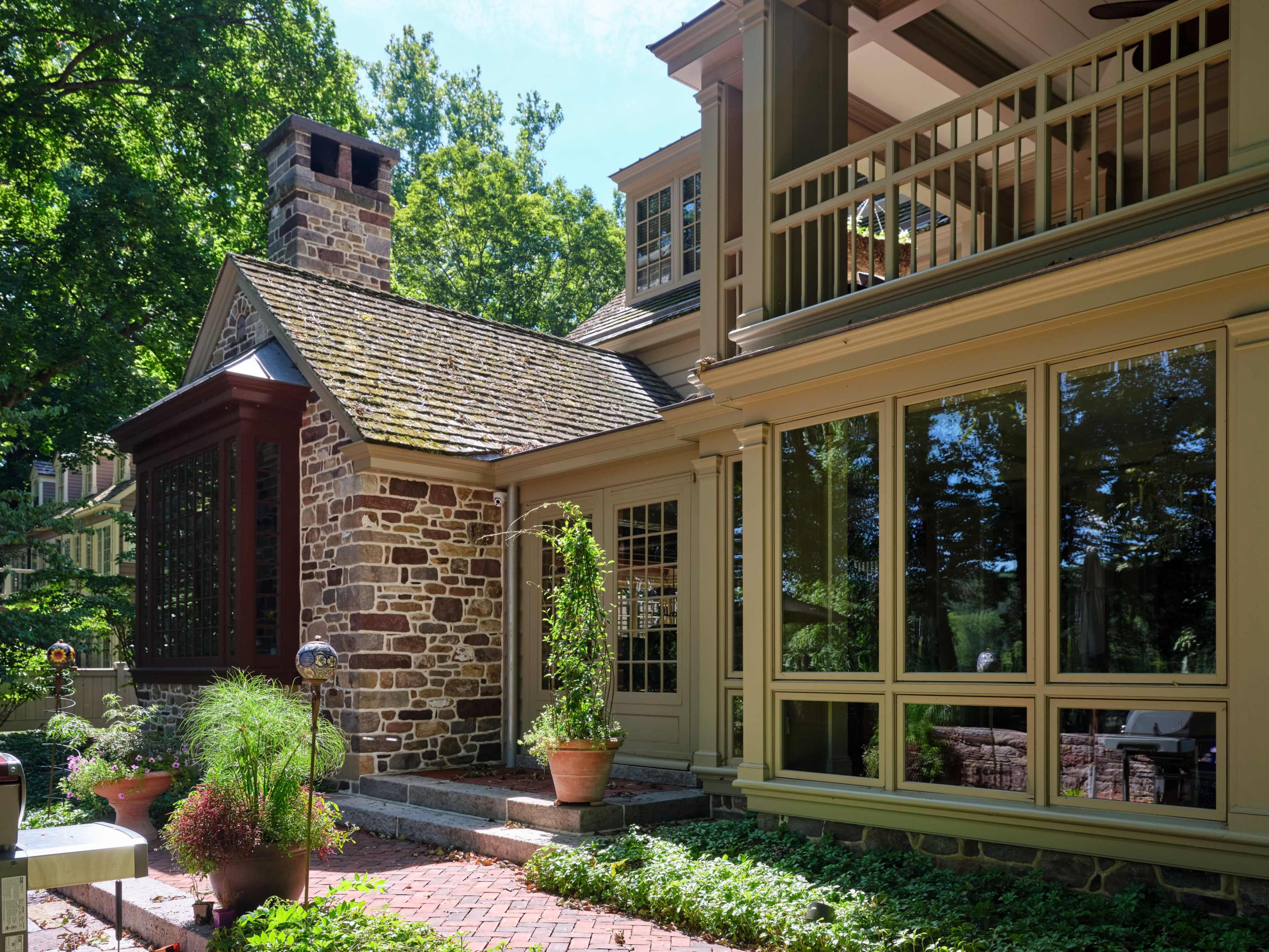 A stone and wood house with large windows, a chimney, and a patio area surrounded by greenery.