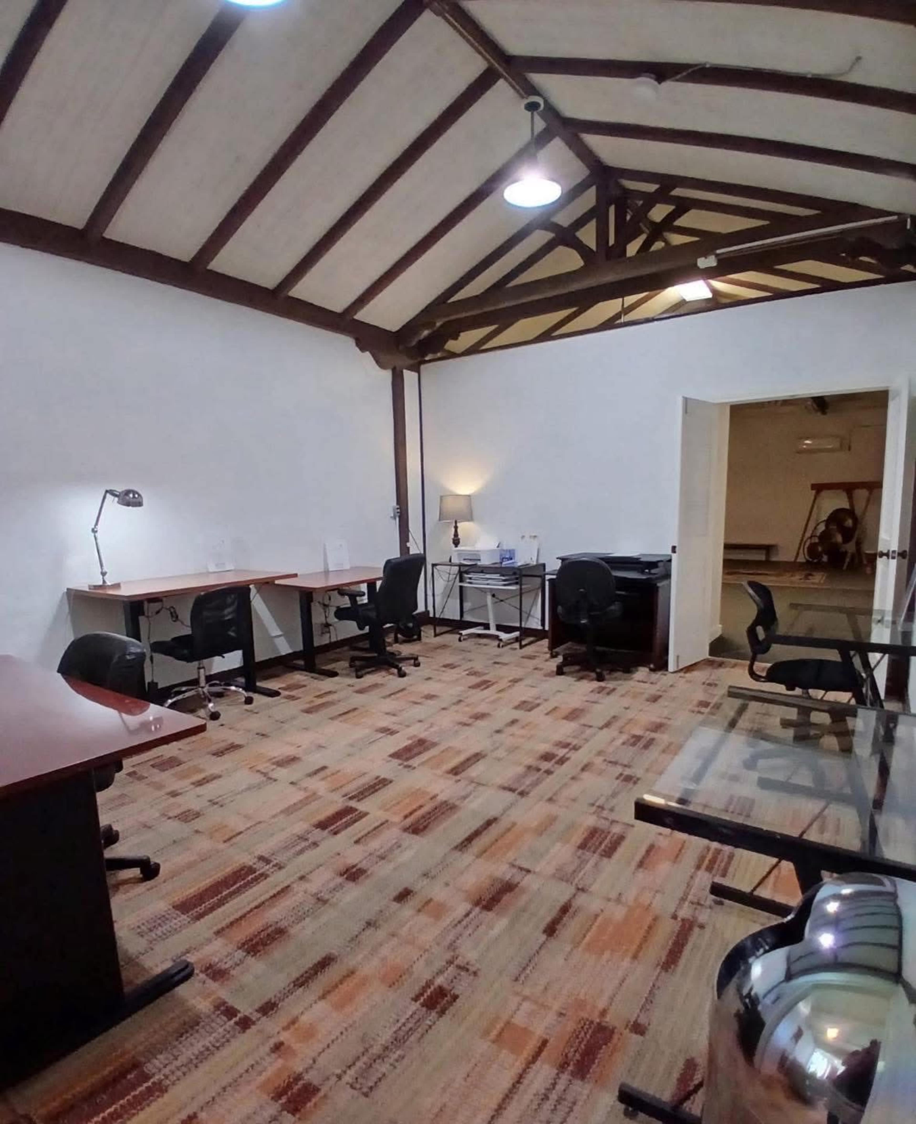 The image shows a spacious, well-lit office with several desks and chairs arranged on a patterned carpet, featuring exposed wooden beams on the ceiling.