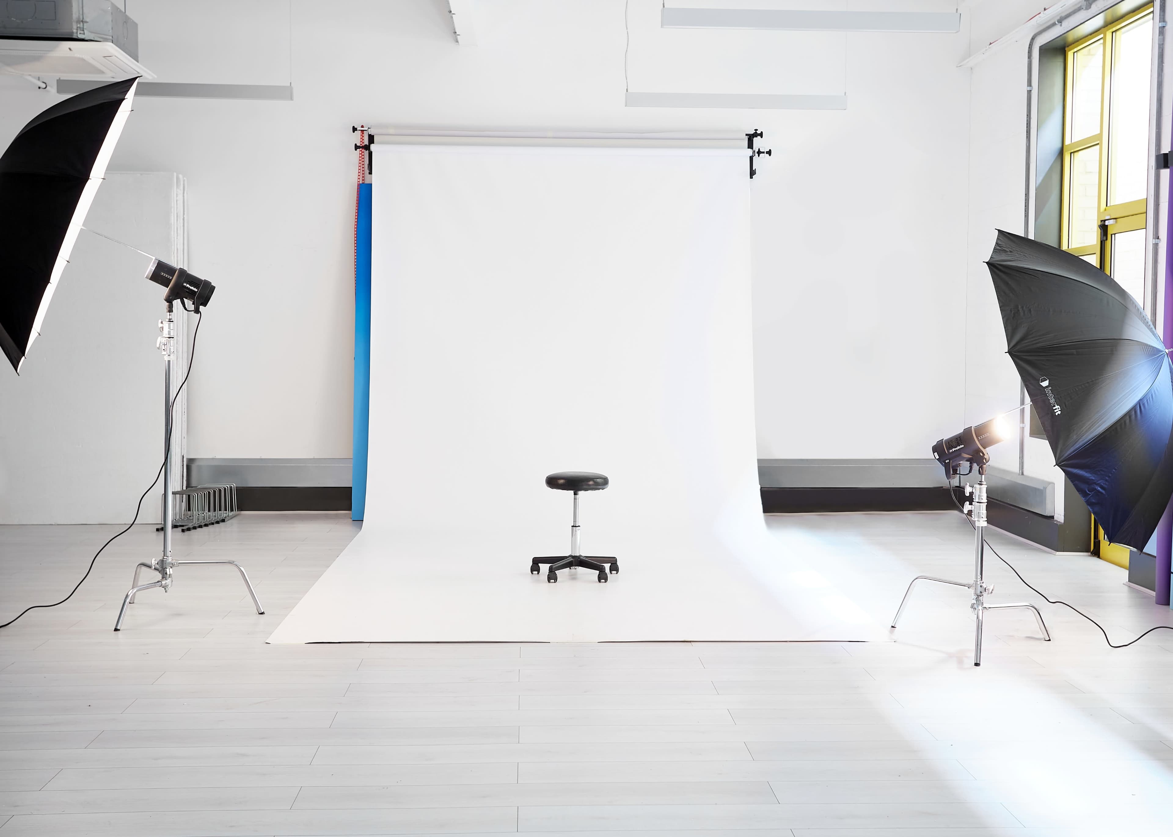 A photography studio features a white backdrop with a stool in the center, illuminated by two softbox lights.