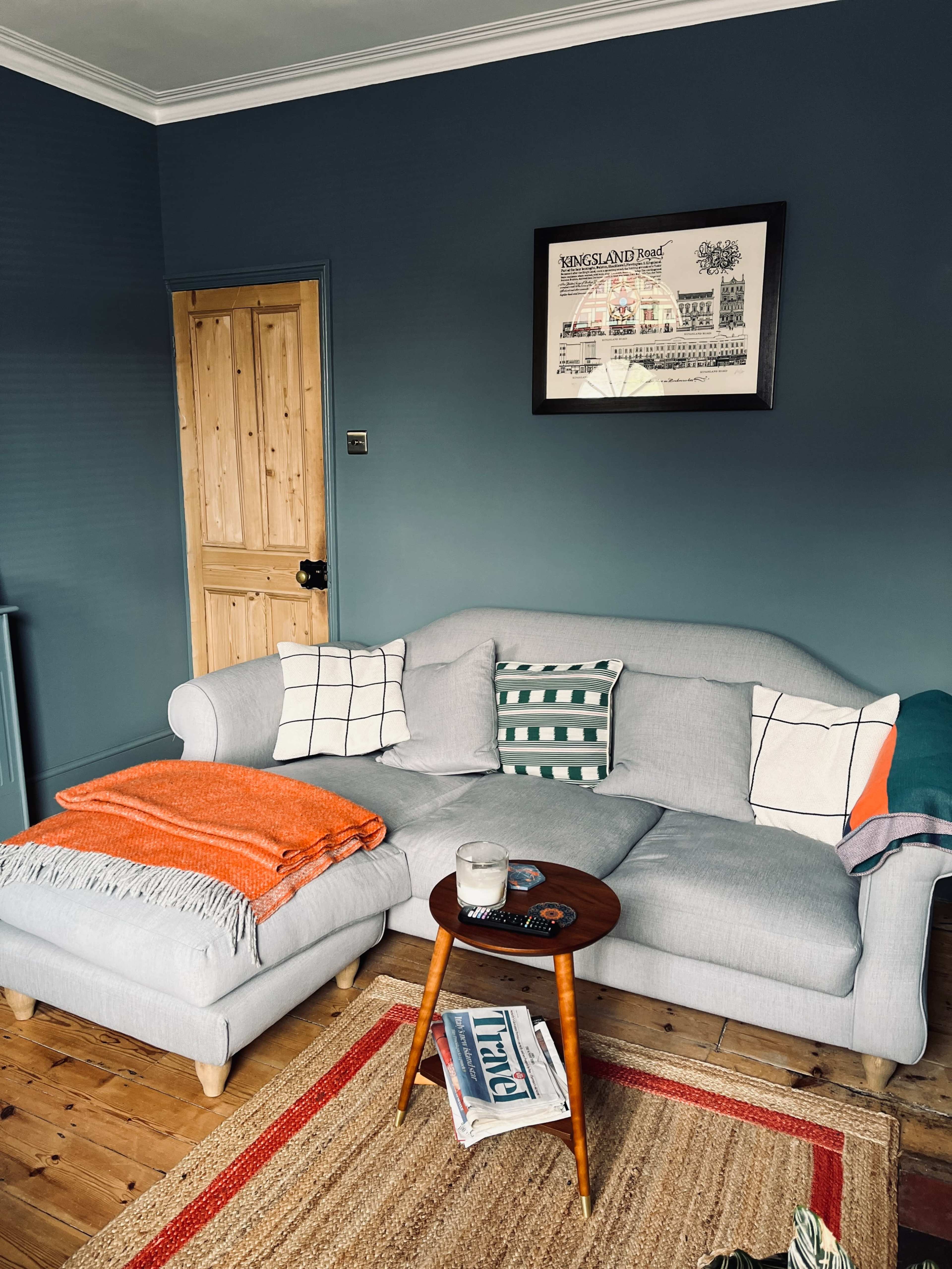 The living room features a gray sofa with patterned cushions, an orange throw blanket, a wooden side table, and a textured rug on a wooden floor.
