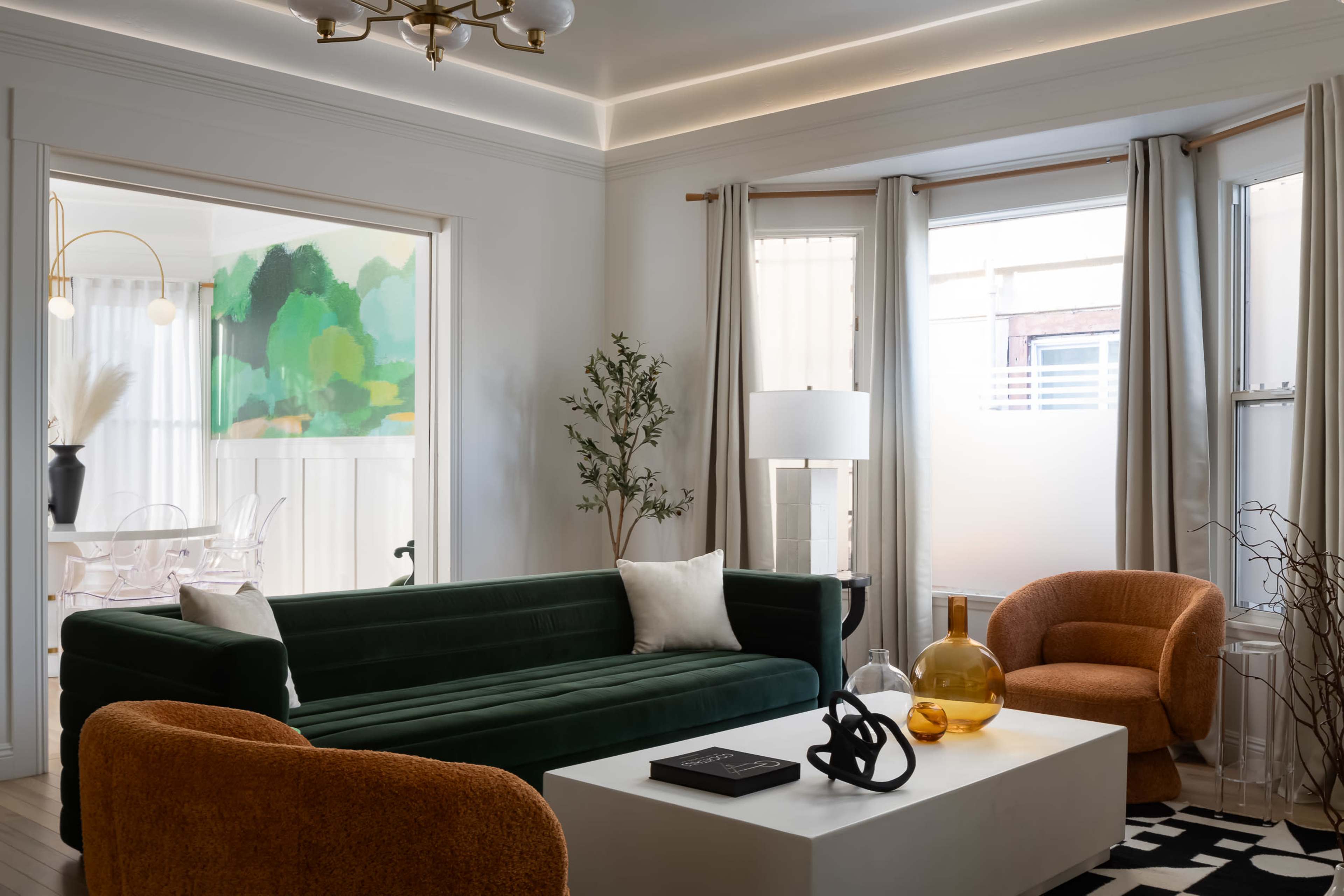 The living room features a green velvet sofa, two orange chairs, a white coffee table, and large windows with sheer curtains, complemented by a large abstract painting in the adjacent dining area.
