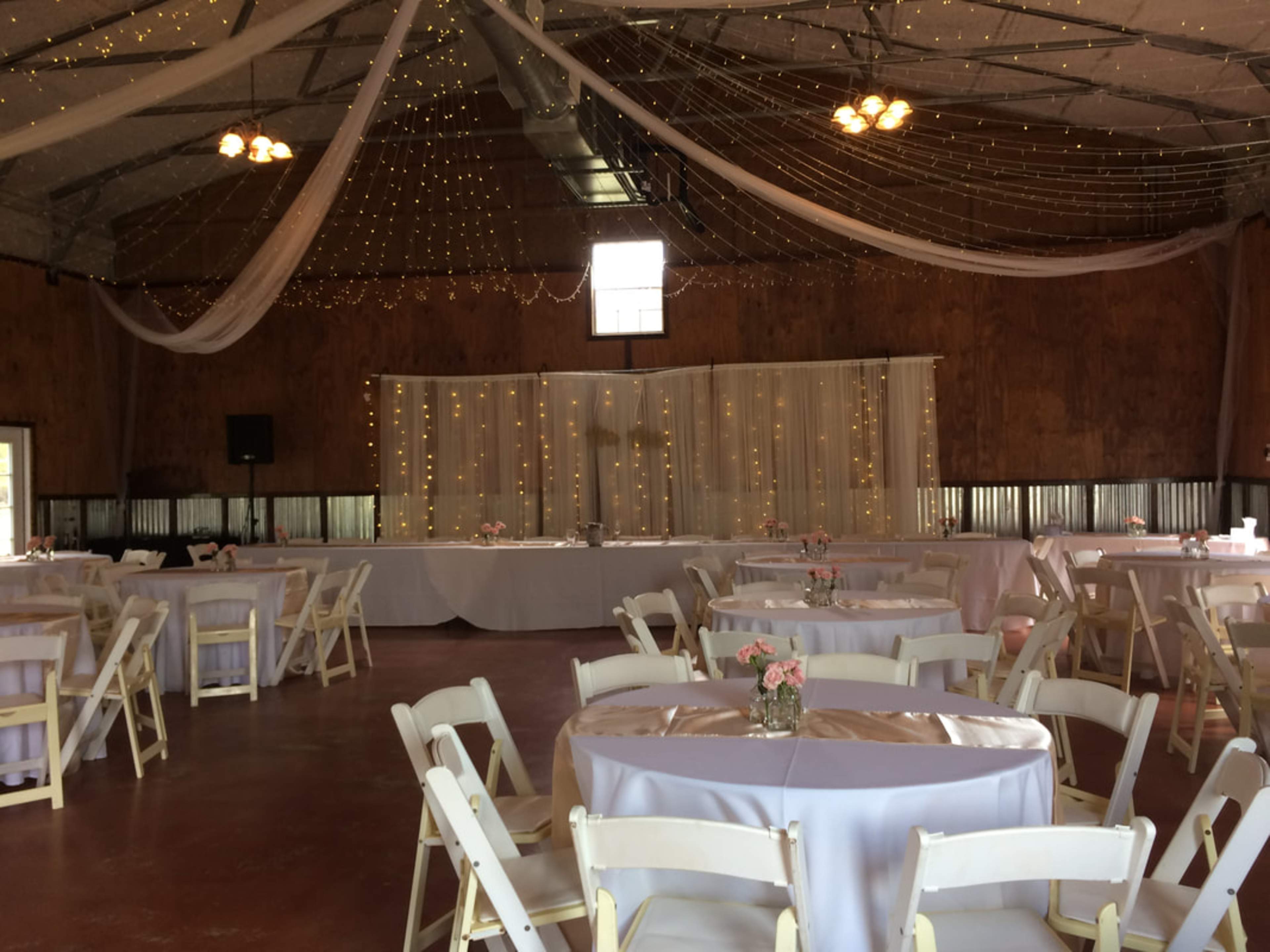 The image shows a rustic event space decorated with string lights and white drapery, featuring round tables set for a gathering.