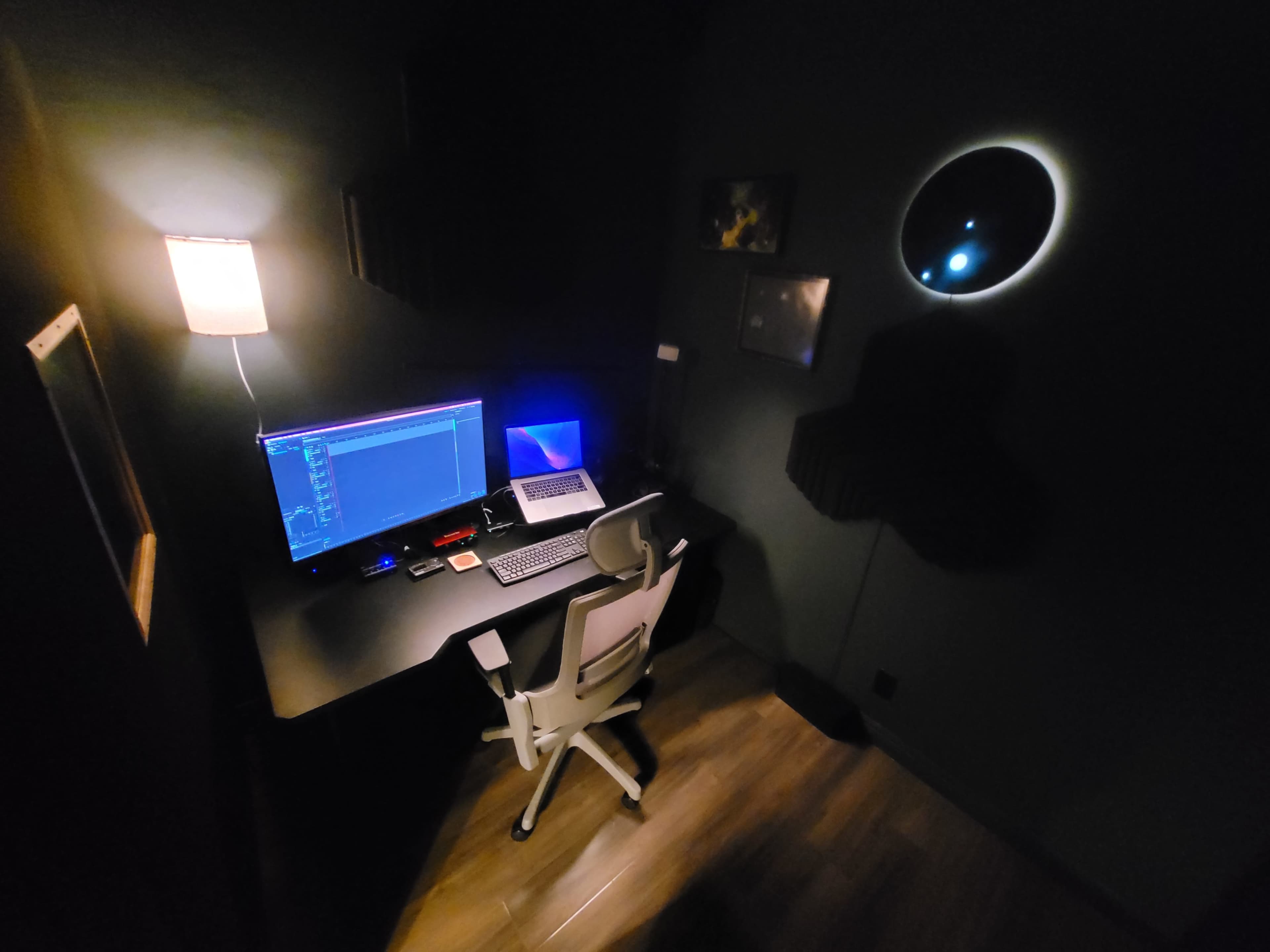 A dimly lit home office features a desk with a computer, a lamp, and a circular window casting light on the wall.