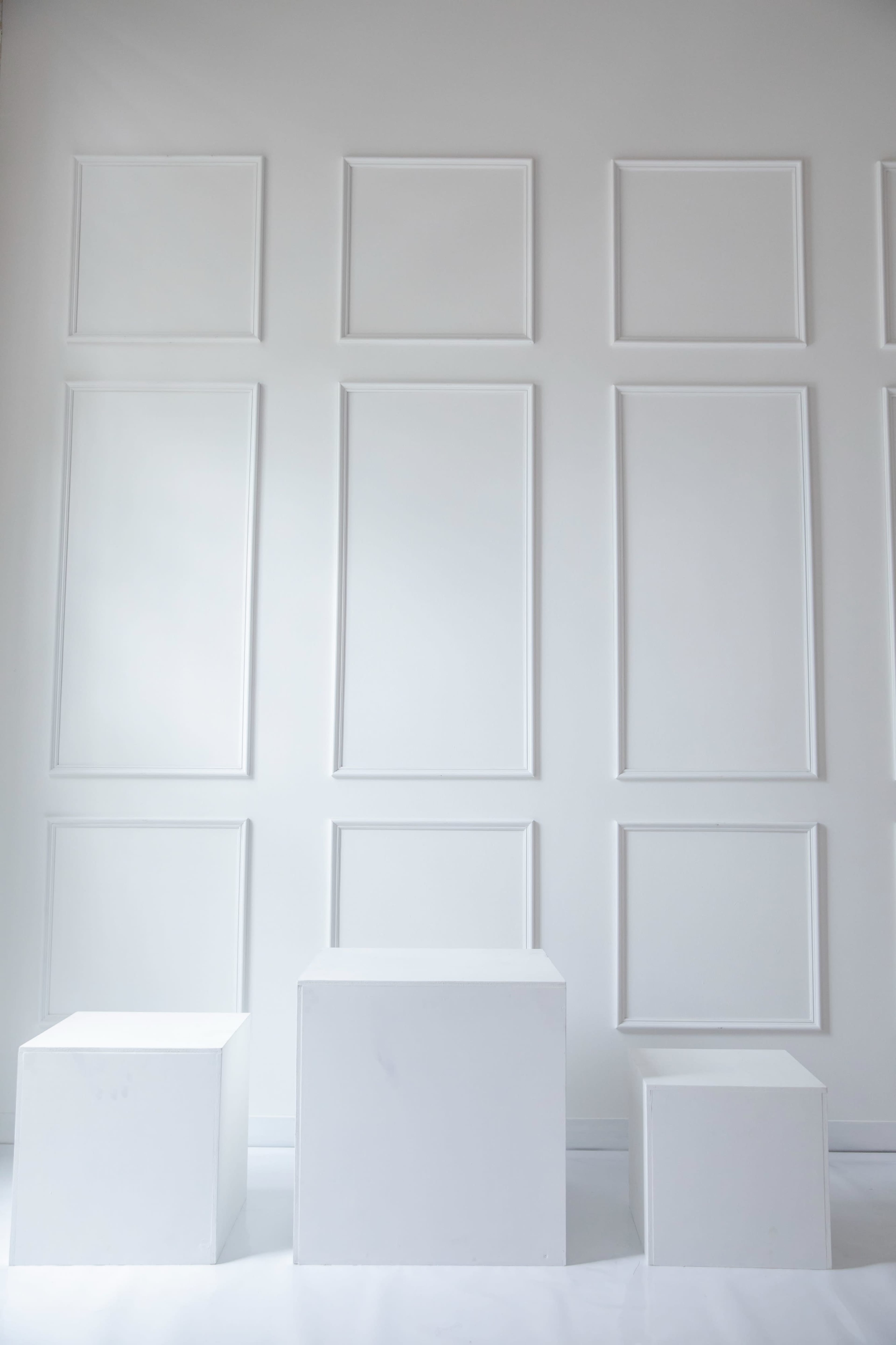 The image features three white geometric pedestals with varying heights positioned against a plain white wall adorned with rectangular frames.