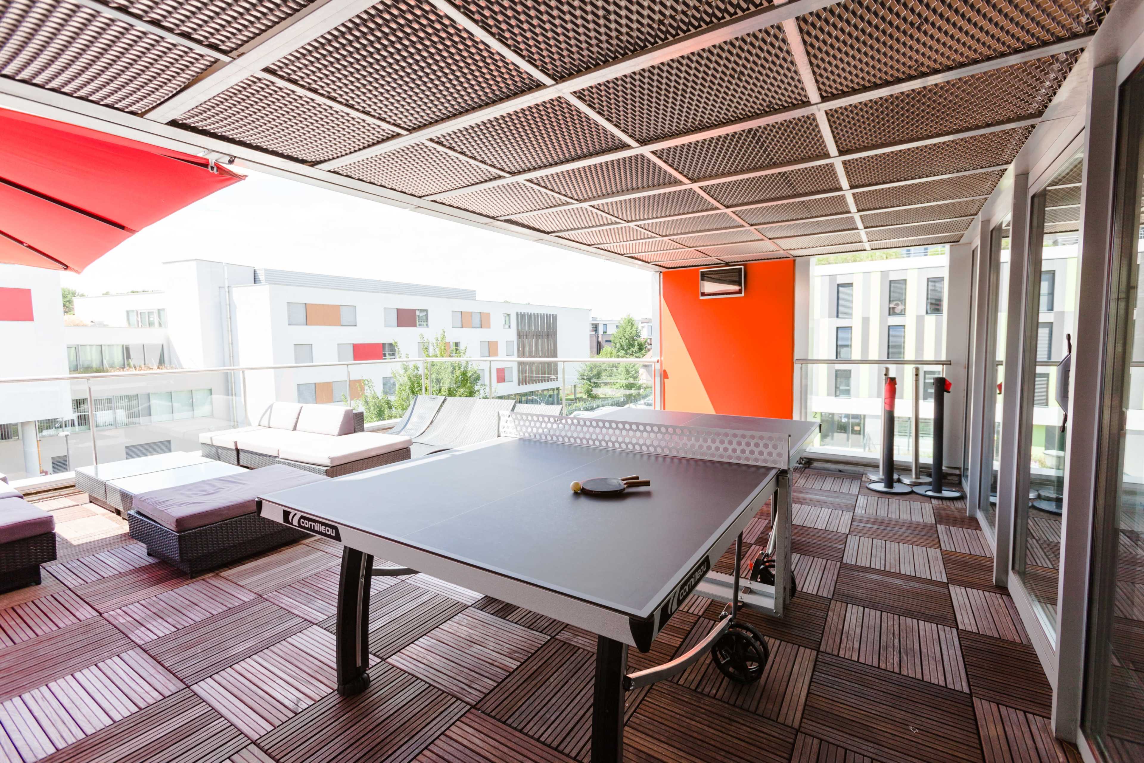 The image shows a modern balcony with a table tennis setup, surrounded by lounge chairs and buildings in the background.