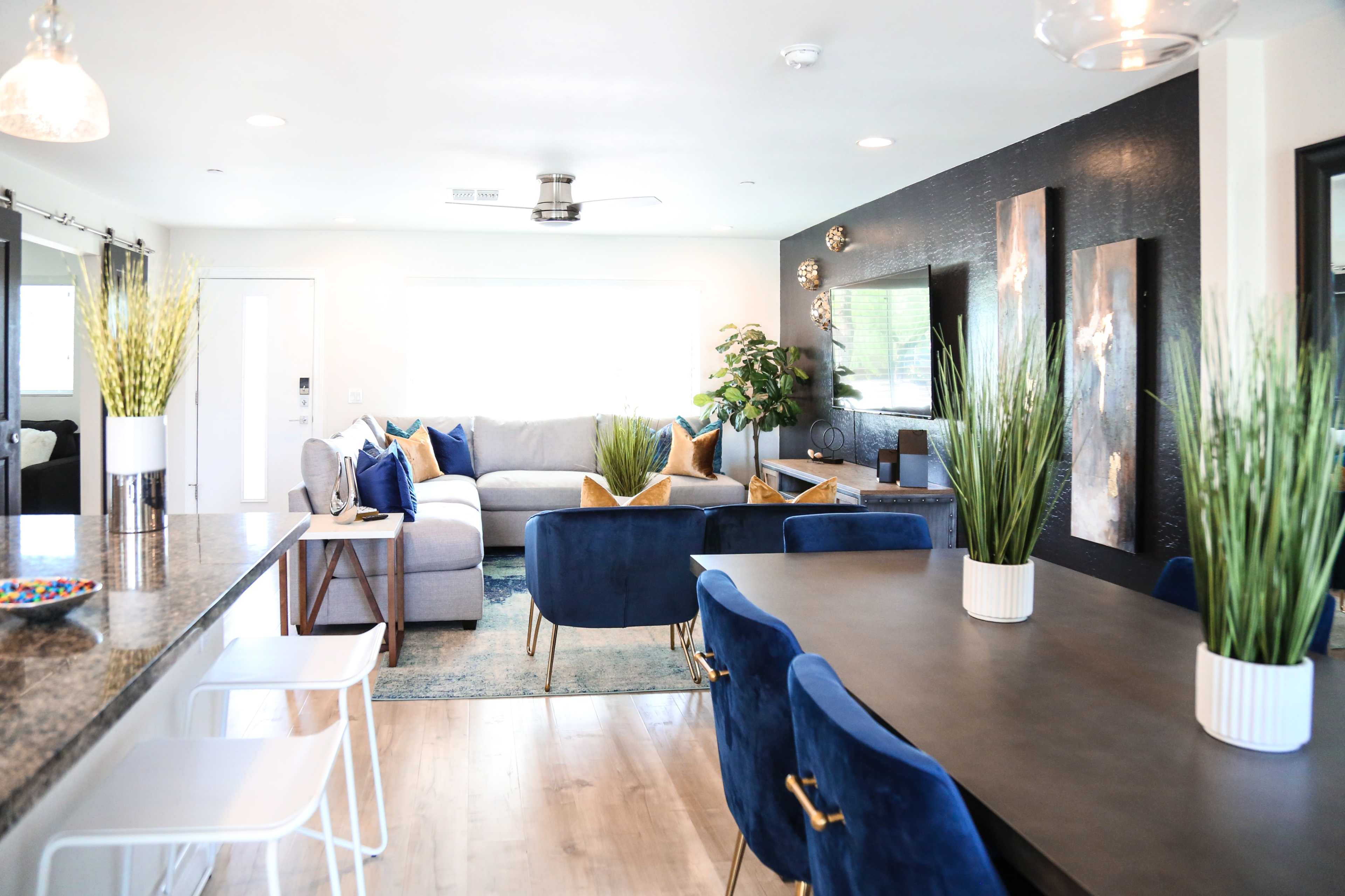 A modern living space features a gray sofa with colorful pillows, a dining area with a dark table and blue chairs, and decorative plants throughout the room.