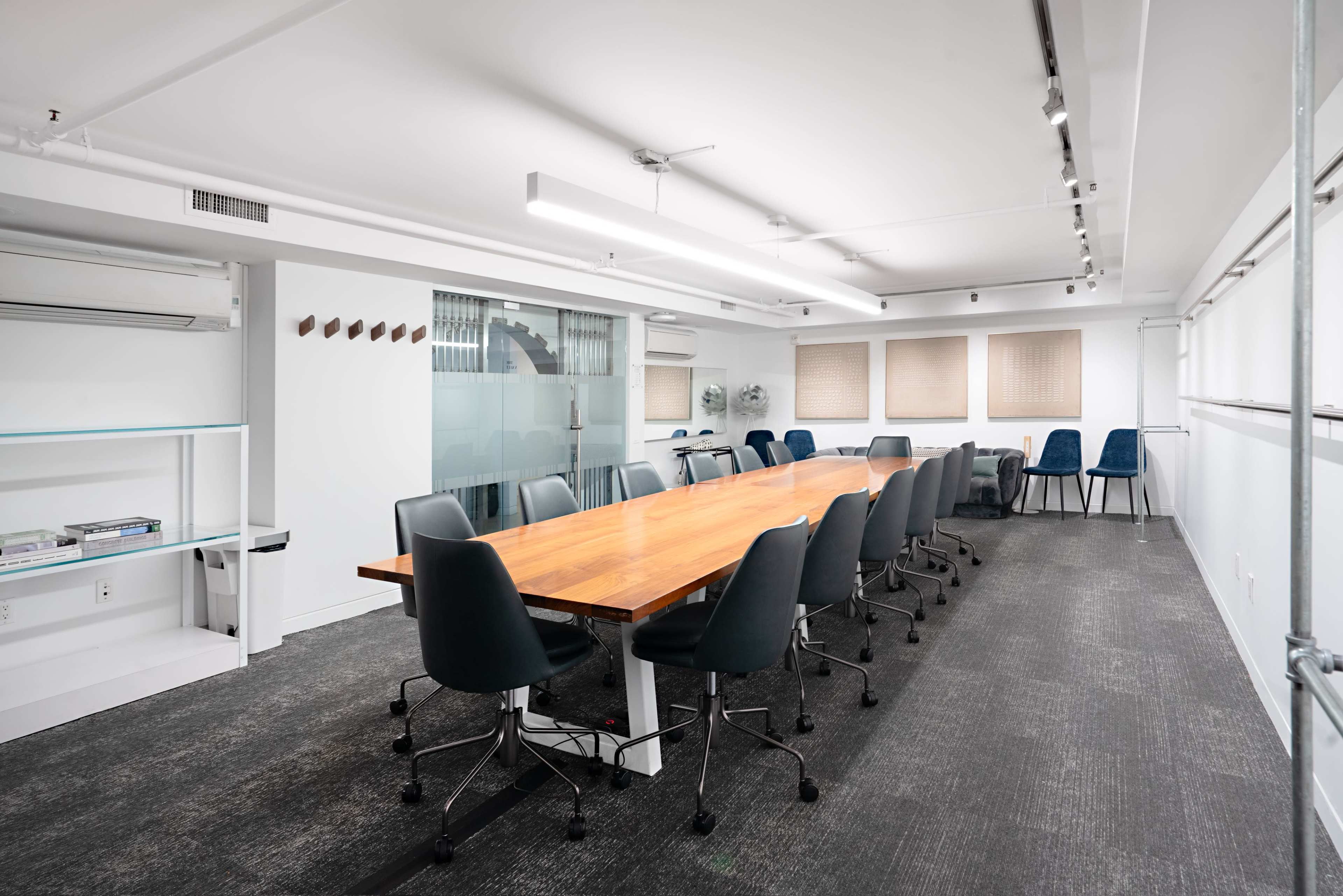 A long wooden table surrounded by rolling chairs fills a modern, well-lit conference room with a clean design and glass partition.