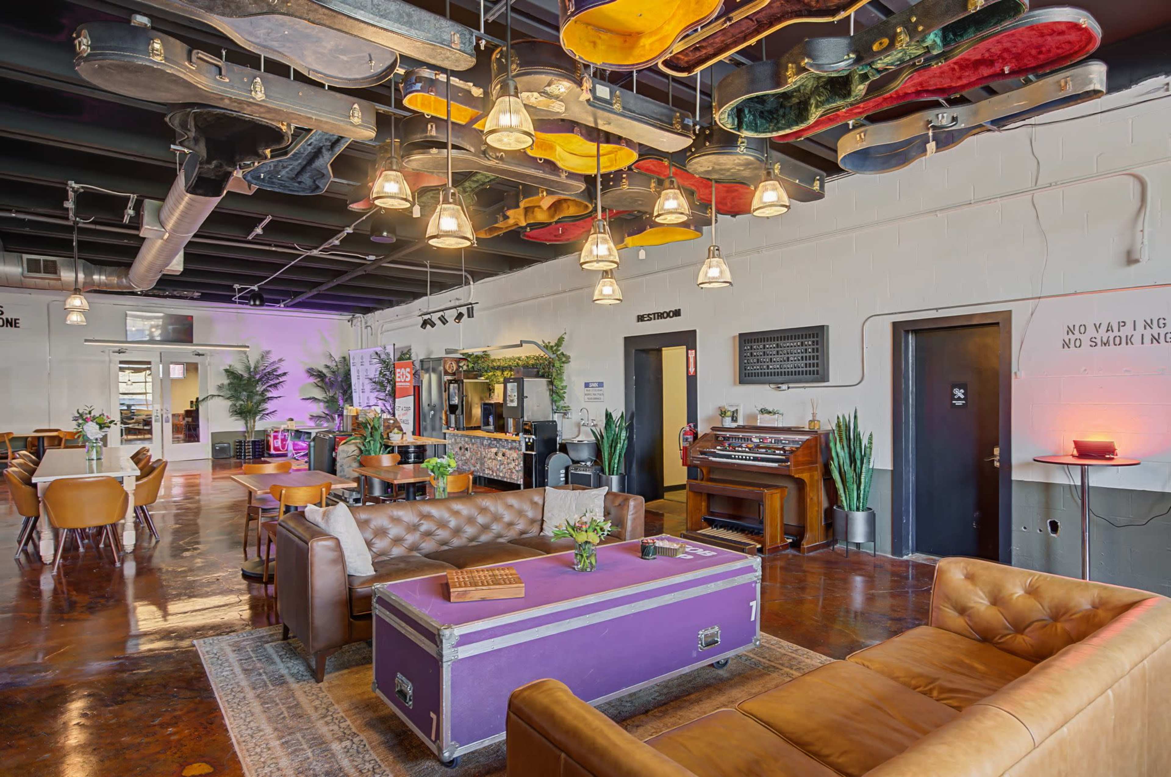 The image features a spacious lounge area with leather seating, a vibrant purple coffee table, and a ceiling adorned with various guitars, surrounded by plants and soft lighting.