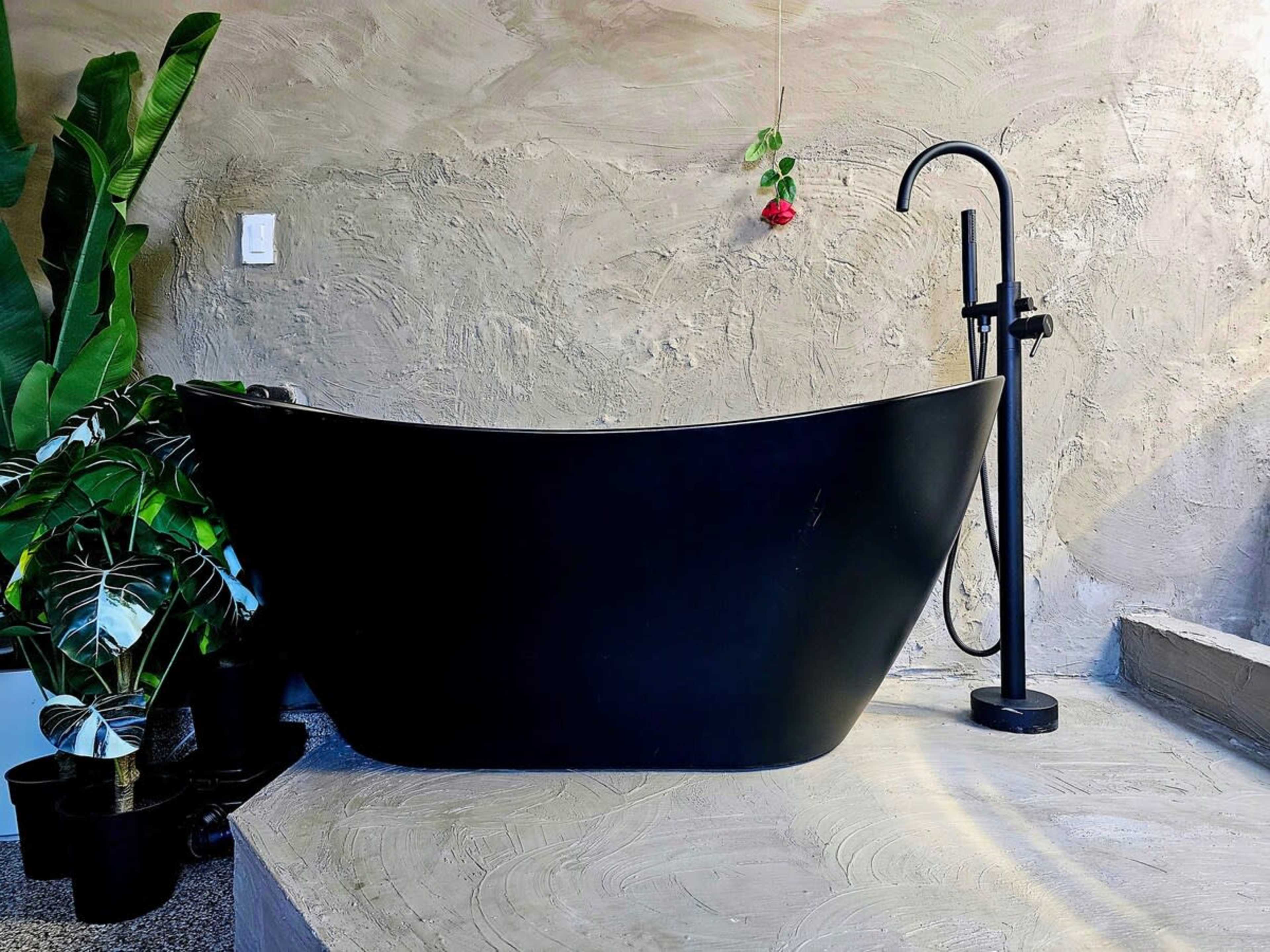 The image shows a modern black bathtub on a textured concrete surface, accompanied by a floor-mounted faucet and greenery in the background.
