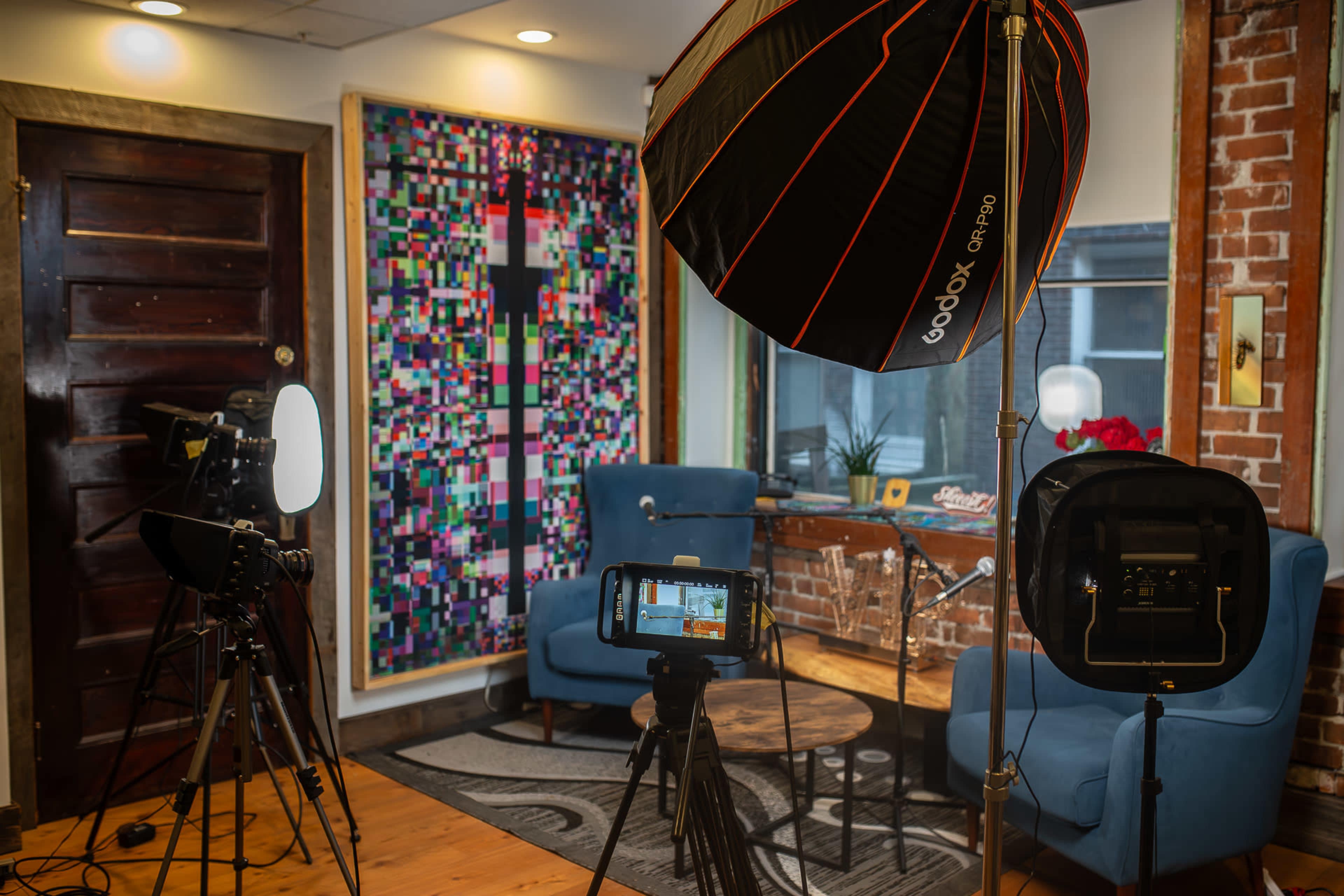 The image shows a small indoor studio setup featuring two blue chairs, various lighting equipment, a camera on a tripod, and a colorful abstract artwork on the wall.