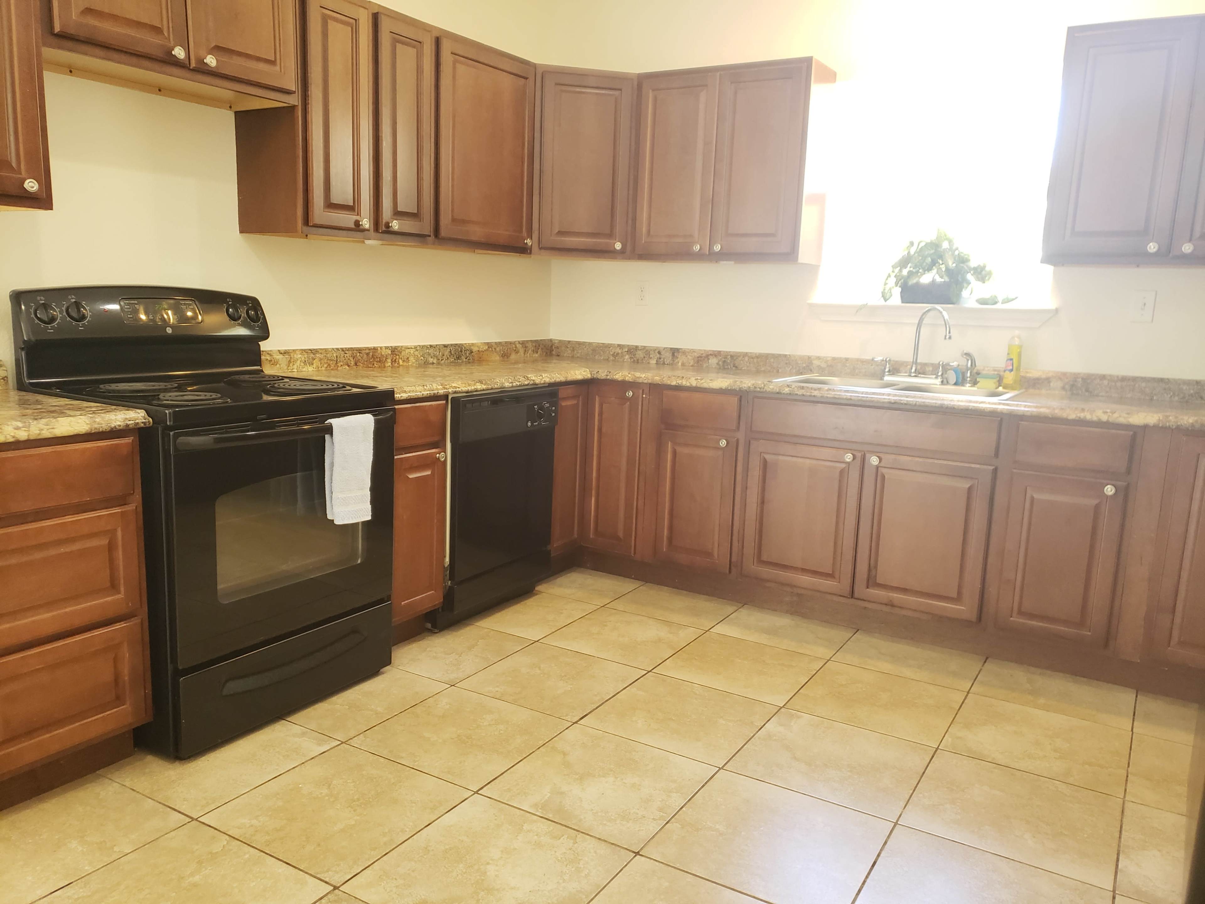A kitchen with dark wooden cabinets, a black stove, a black dishwasher, and a sink under a window, with beige tiled flooring.