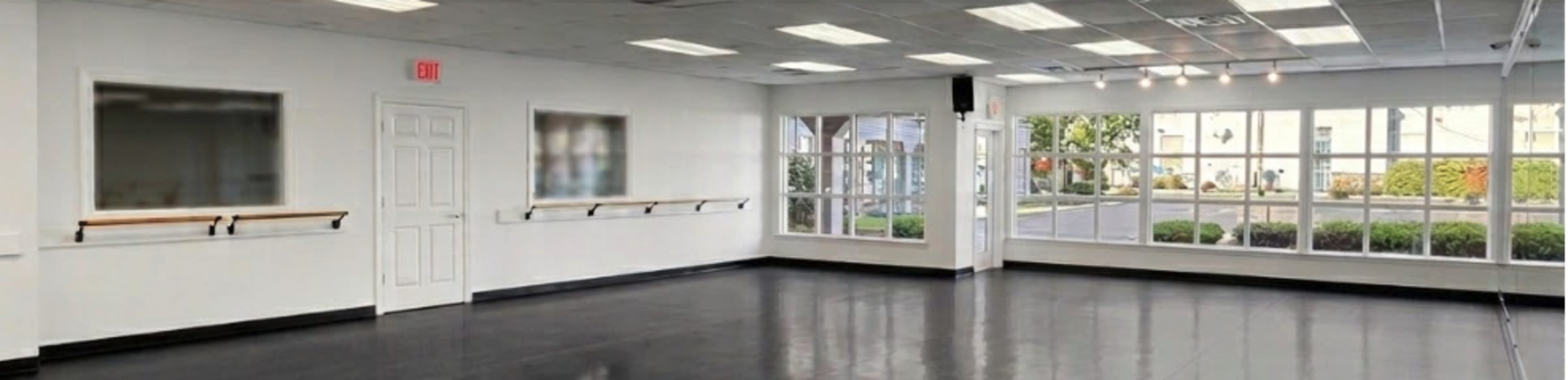The image shows a bright, empty dance studio with large windows and ballet barres mounted on the walls.