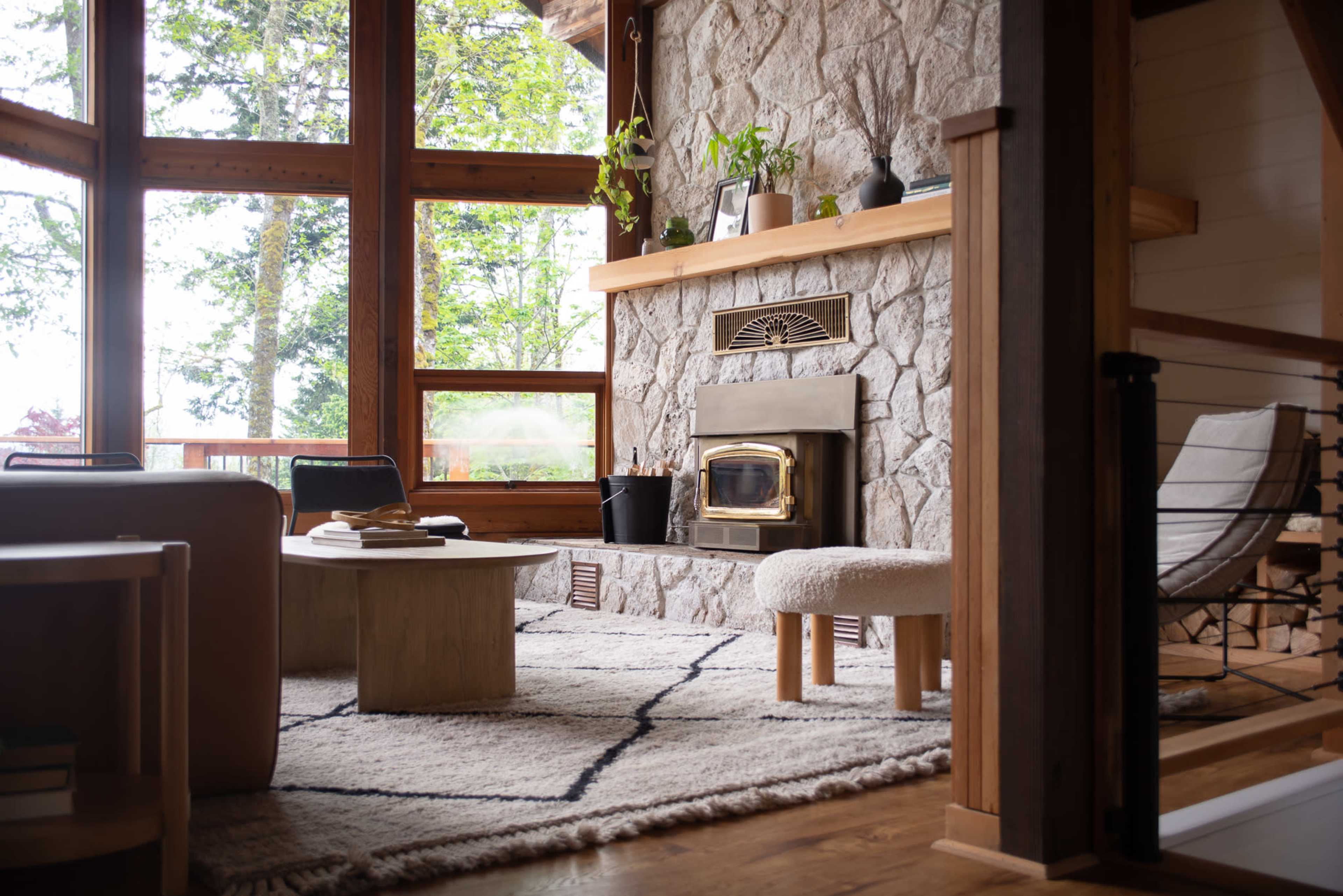 A cozy living room features a stone wall with a fireplace, large windows with views of trees, and minimalist furniture arranged around a coffee table.