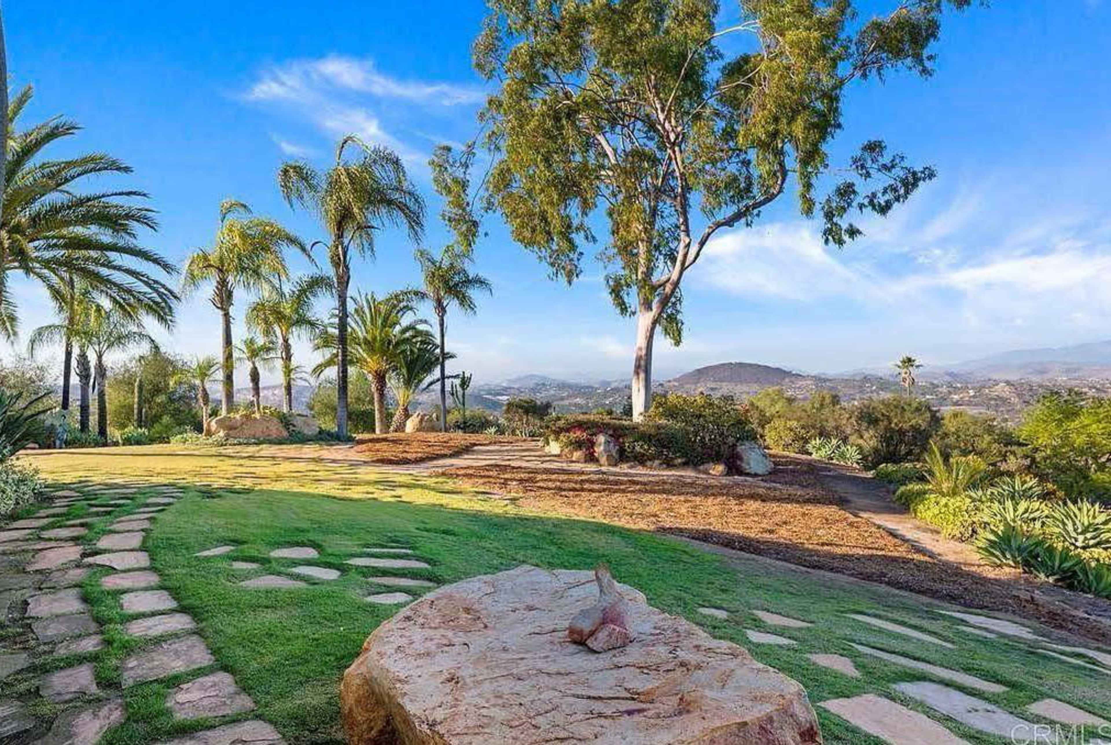 A landscaped backyard with stone pathways, palm trees, and a view of distant hills under a blue sky.