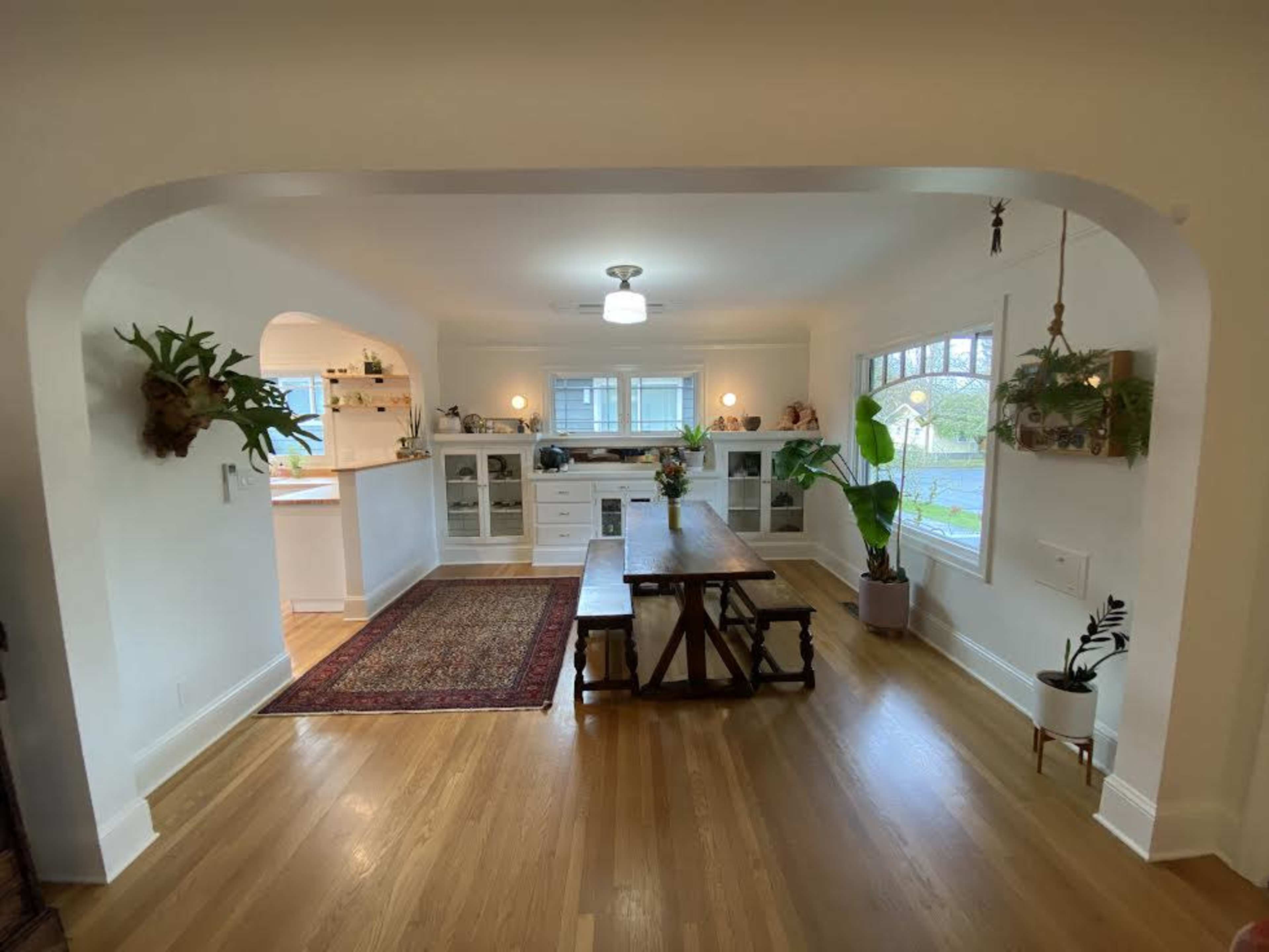 A dining area features a long wooden table, surrounded by plants and a rug, opening into a well-lit kitchen space with white cabinetry.