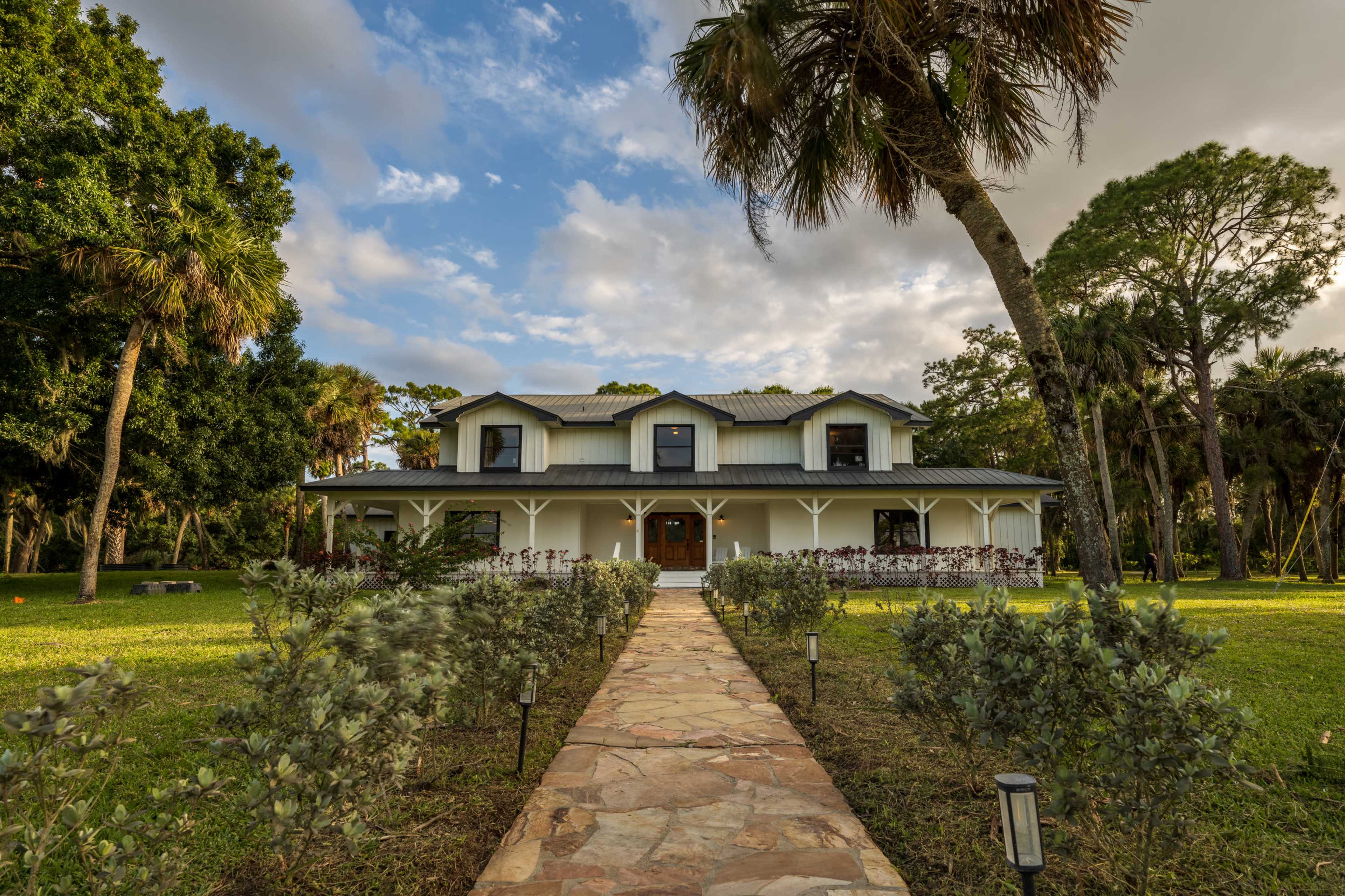 A two-story white house with a metal roof is surrounded by greenery and a stone pathway leading to the front entrance.